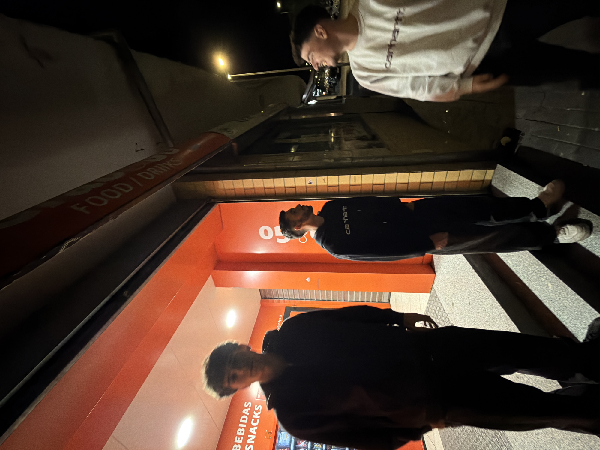 Three young men stand outside a vending machine store at night. The store has orange walls with "BEBIDAS SNACKS" and "FOOD / DRINKS" signs. The man closest to the camera is wearing a dark-colored hooded sweatshirt. The next man is also wearing a dark-colored sweatshirt. The third man is wearing a light-colored sweatshirt with "Carhartt" written on it. The location is likely in the city of Bragança, Portugal.