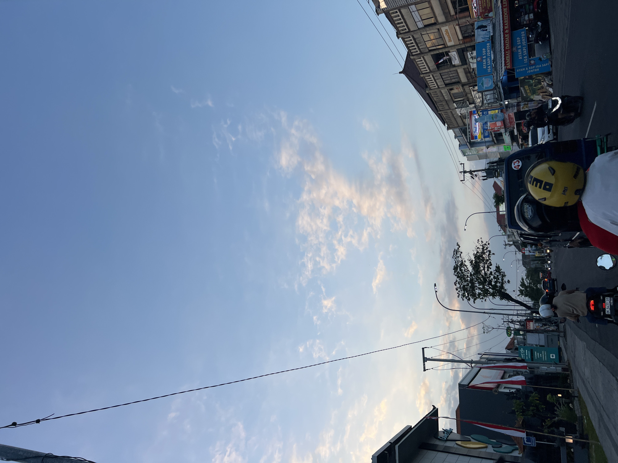 The image depicts a street scene in Kuta, Indonesia, during twilight. A clear blue sky with scattered wispy clouds dominates the upper portion. In the foreground, two individuals are riding a motorcycle, with the person in the rear wearing a yellow helmet. To the left, a building with flags displaying the Indonesian national colors (red and white) is visible. The street itself is lined with businesses, including shops and restaurants, indicated by various signs. Traffic, consisting of other motorcycles and cars, is present on the road, suggesting a moderately busy urban environment. The overall atmosphere is characteristic of a late afternoon or early evening in a bustling Indonesian city.