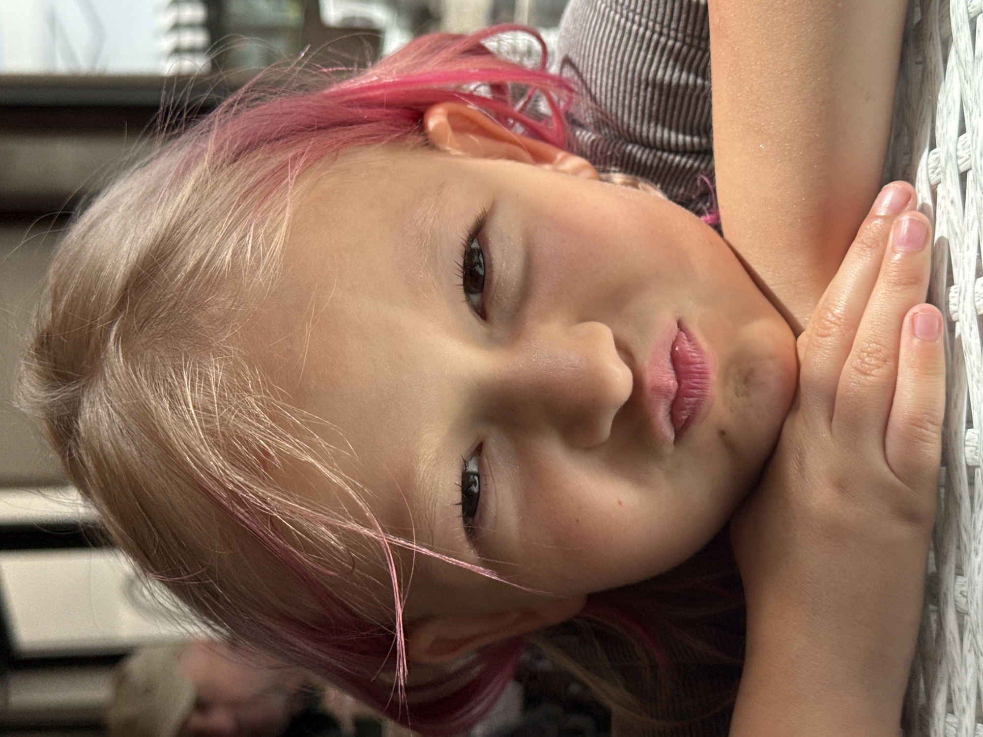 The close-up, portrait-oriented image shows a young girl with light blonde hair and prominent bright pink streaks. She is resting her head on her right hand and arm, which are propped against a white, woven or wicker-like surface, possibly a chair or fence. The image is rotated 90 degrees counter-clockwise, making her head appear tilted to the right.
The girl is the primary subject, looking directly at the viewer with open, dark eyes and long eyelashes. Her lips are slightly pursed, giving her a contemplative or subtly pouty expression. Her skin is fair, with some natural texture visible. Her fingers and fingernails, which are short and natural, are visible on her propped hand. She is wearing a garment with thin vertical stripes, visible on her upper right arm and shoulder.
The background is softly blurred, showing indistinct shapes that suggest an indoor or shaded outdoor setting. A very blurry figure, possibly an adult, is visible in the lower left background. The lighting appears to be soft and even, indicating daytime but not harsh direct sunlight. There are no specific location cues or visible text. The overall impression is a casual, intimate moment.
