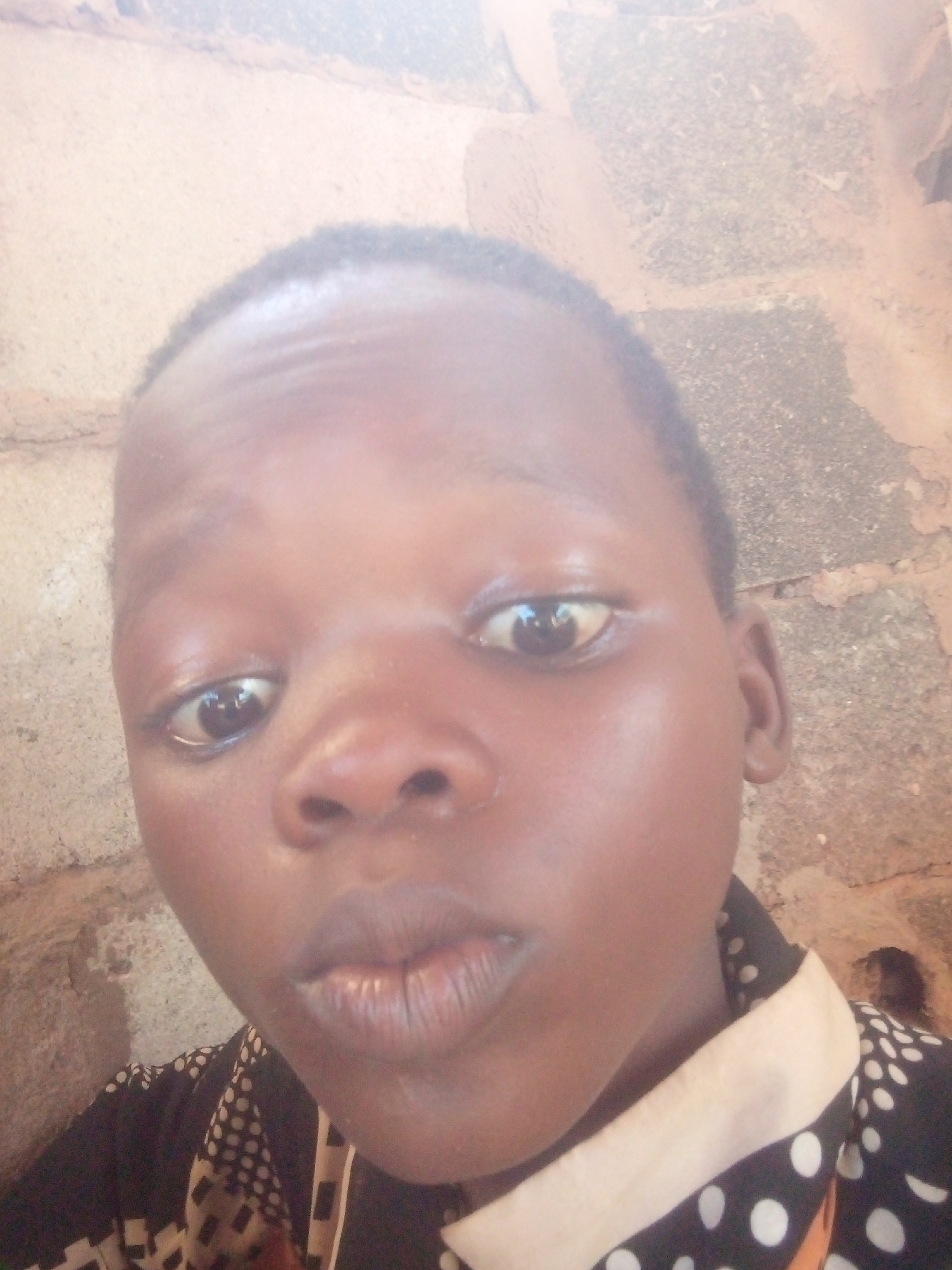 A close-up shot captures a dark-skinned individual with short hair. They are positioned against a wall composed of brick-like structures, possibly made of mud or clay. The person is wearing a dark shirt with light-colored polka dots.
The city and state are unknown.