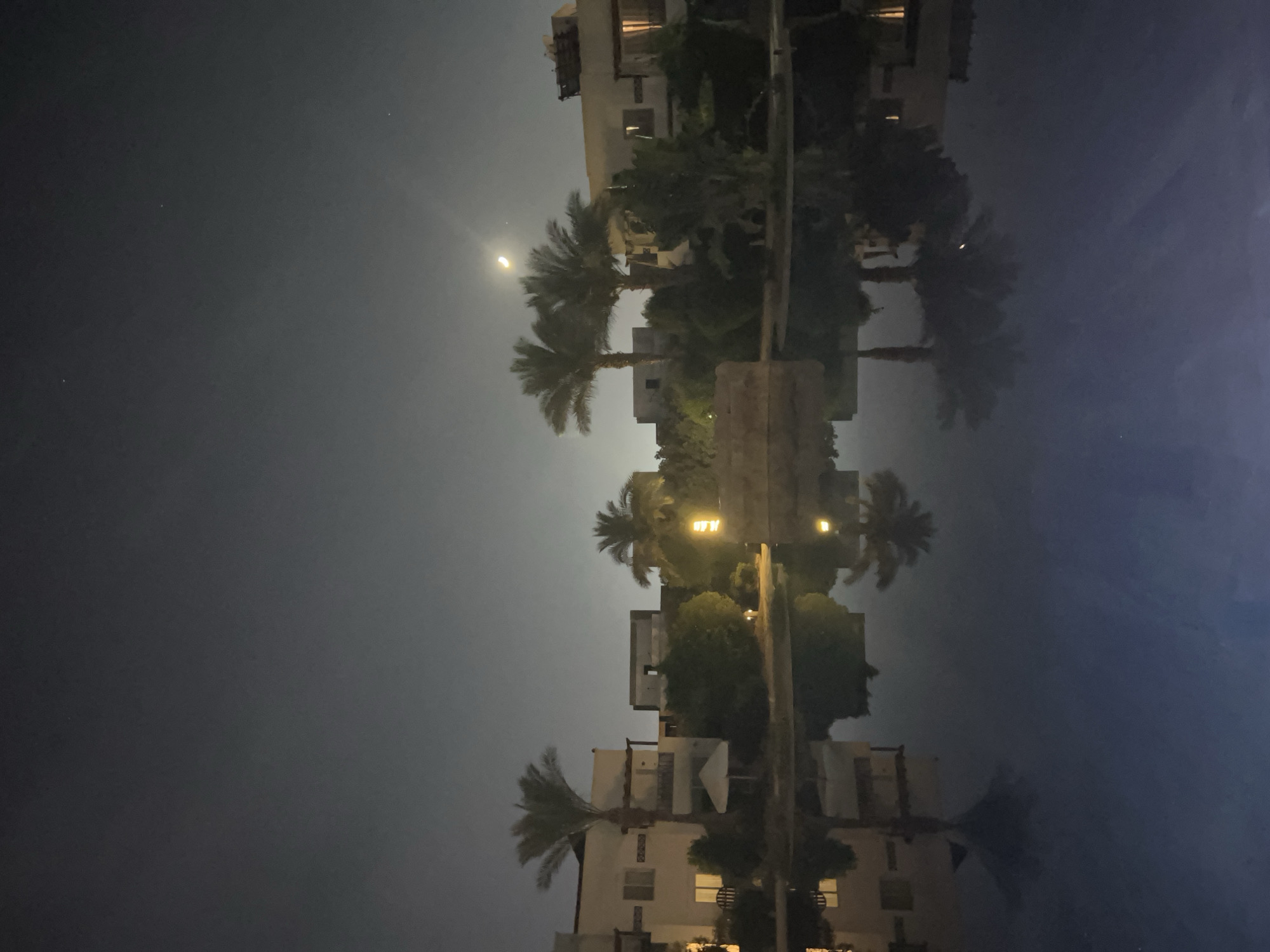 An outdoor night scene in Sharm El Sheikh, Egypt, captures a tranquil view of a resort with illuminated buildings and palm trees reflected in a body of water. The sky is dark with a crescent moon visible, and a few stars are scattered across it. The architecture suggests a modern resort style, with light-colored buildings and balconies. The water in the foreground, possibly a pool or a calm sea, creates a clear reflection of the scene above, doubling the visual impact of the illuminated structures and vegetation. The overall atmosphere is serene and peaceful, typical of a vacation destination at night.