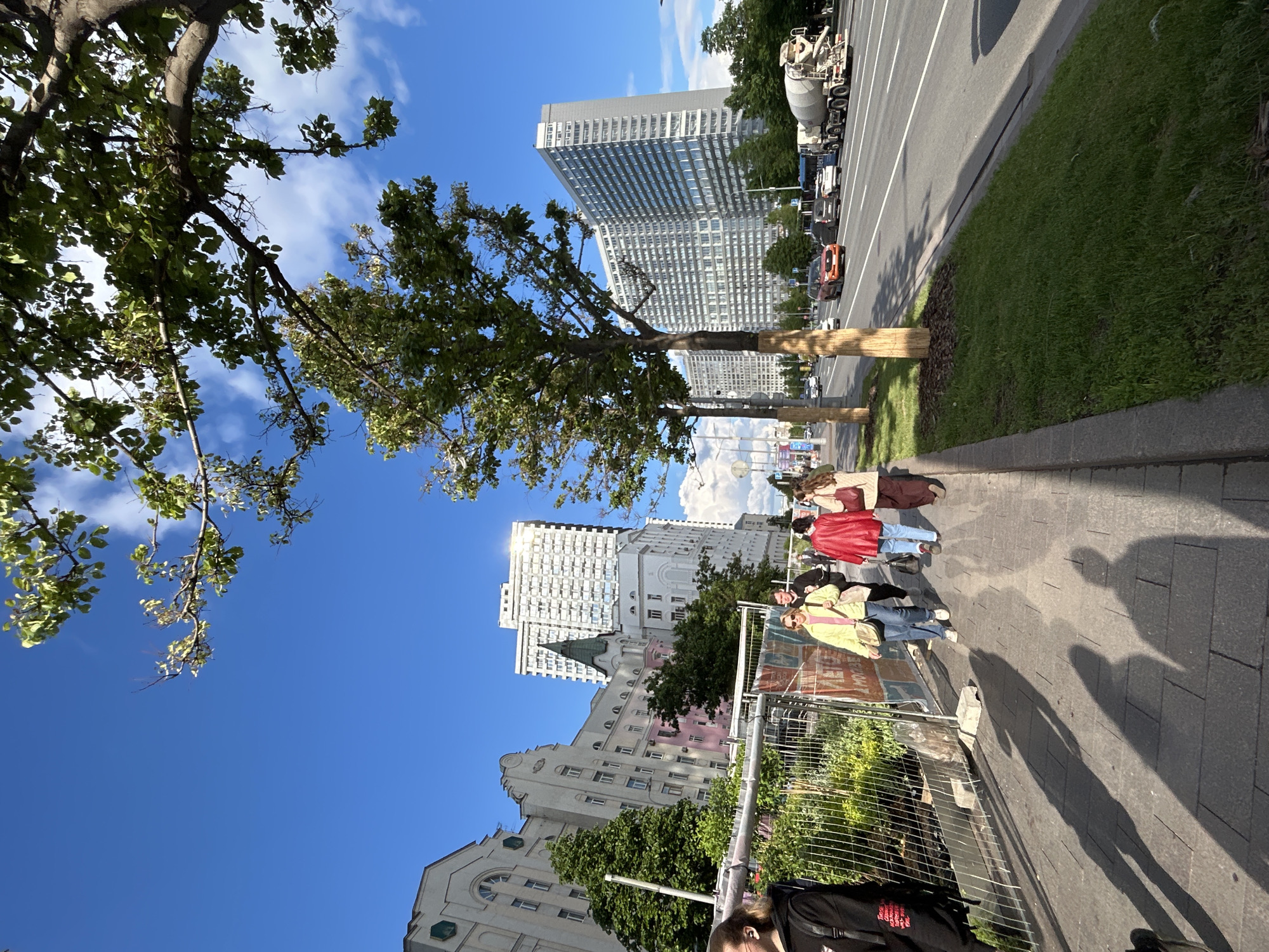 A street scene showing a sidewalk with pedestrians walking along it. The sidewalk is paved with rectangular stones. A temporary fence is visible near the foreground, running parallel to the sidewalk. On the other side of the fence, there are some plants and greenery. In the background, there are multiple buildings of varying architectural styles, including tall modern skyscrapers and older buildings with decorative elements. Trees line the street, providing shade. The sky is blue with scattered clouds. On the right side of the image, a road with vehicles is visible, including a concrete mixer truck.