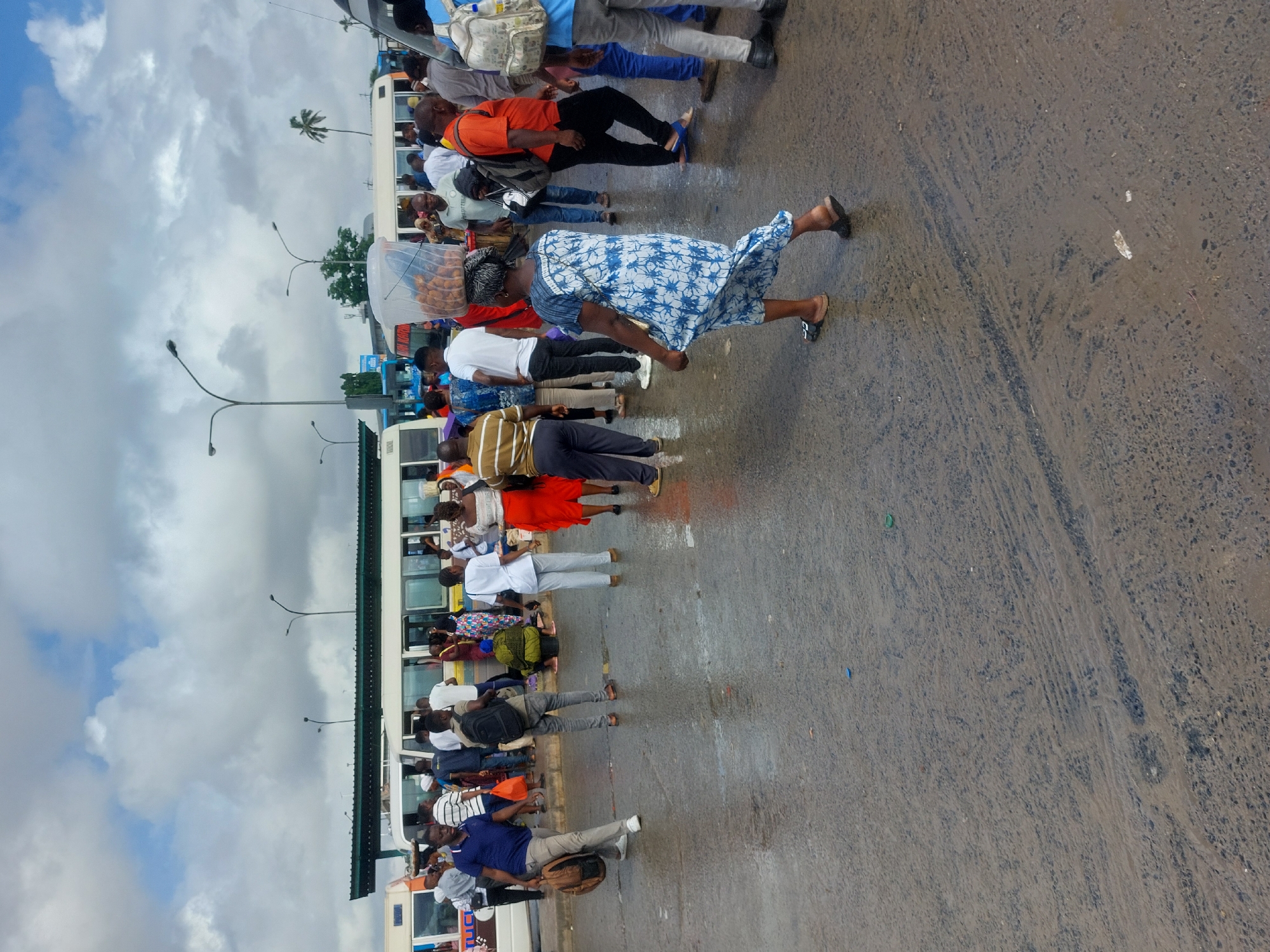 The media file captures a bustling street scene in what appears to be a bus stop or public transport area, consistent with an urban setting like Dar es Salaam, Tanzania. The image is oriented vertically (rotated 90 degrees clockwise).
The scene is dominated by a large crowd of people, primarily adults, many of whom are standing and waiting. Several individuals are seen near or inside white buses, suggesting public transportation is a central activity. One prominent figure in the mid-ground is a woman walking away from the viewer, wearing a blue and white patterned dress and carrying a clear plastic container on her head, likely holding food items like snacks or pastries. Other people are casually dressed, some carrying backpacks or bags.
The ground is an asphalt road, visibly wet and possibly muddy in areas, with textured patterns that could be from water flow or tire marks. Small pieces of litter, including what appear to be colorful plastic bottle caps, are scattered on the damp surface. The sky is overcast with thick, grey clouds, indicating a cloudy day, possibly after rain. In the distance, against the cloudy sky, several streetlights with curved arms are visible, as well as the silhouette of a single palm tree. No legible text is clearly visible on any signs or vehicles.
The overall impression is one of everyday urban activity and commute, with people going about their routines in a busy public space.