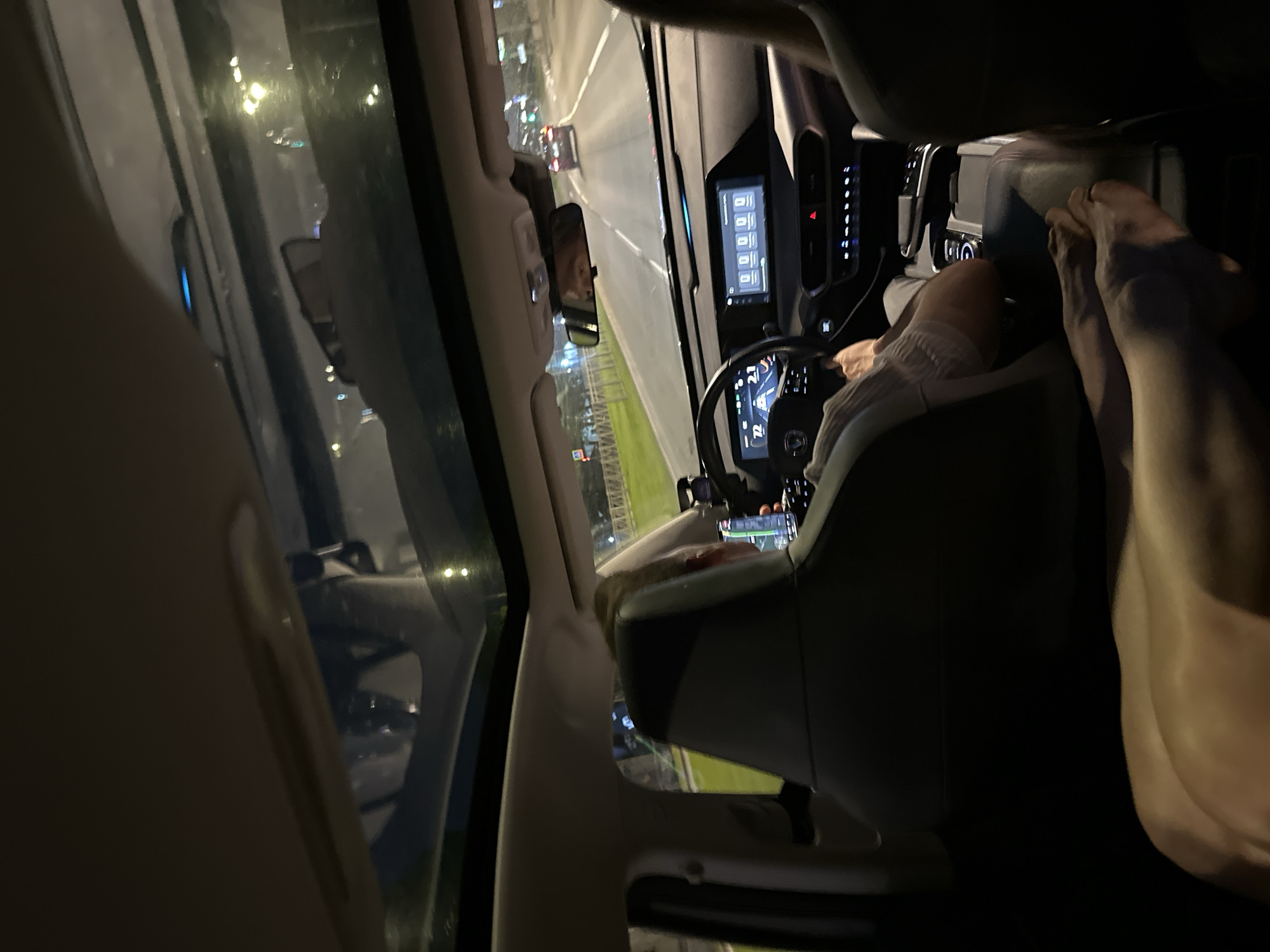 The image shows the interior of a car driving at night on a city street, likely in Saint Petersburg, Russia, given the context. The driver, a person with blonde hair, is visible from behind, operating the vehicle. The dashboard displays show the car's speed and navigation information. In the foreground, another person's legs and feet are stretched out, suggesting they are a passenger in the back seat. The car has a panoramic sunroof, through which city lights and streetlights are visible, illuminating the road ahead. The overall scene depicts a typical nighttime drive through an urban environment.