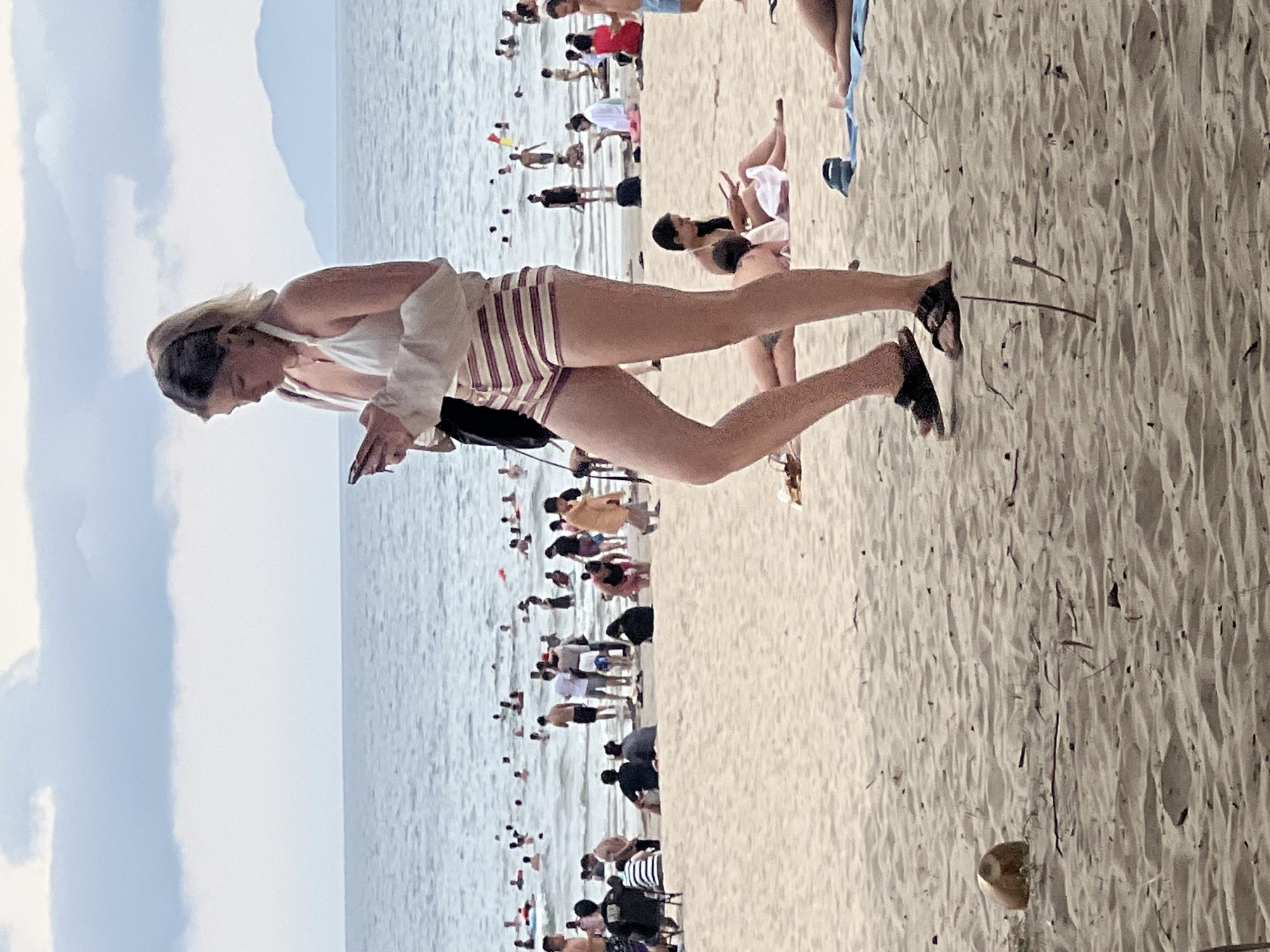 A young woman, wearing a white halter bikini top and striped shorts, stands on a sandy beach, looking down at her phone. Her hair is pulled back in a ponytail. She is wearing sandals and has a light-colored wrap or sweater draped over her arms. In the background, the ocean is visible with many people swimming and relaxing. Further in the distance, mountains can be seen. The scene appears to be on a cloudy day. The image is taken from a slightly low angle. It is likely a beach in Da Nang, Vietnam, given the presence of the ocean, sandy beach, and distant mountains, which are characteristic of the region.