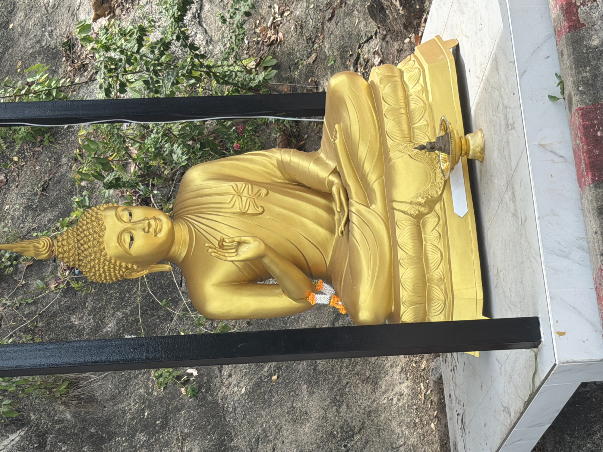 The image shows a golden Buddha statue sitting in a meditative pose on a tiered pedestal. The statue is adorned with a garland of white and orange flowers around its neck. In front of the large statue, on the same platform, is a smaller Buddha figurine placed on a golden offering dish. The platform itself is made of white tiles with a marble-like pattern and is bordered by a concrete curb with red and white stripes. The statue is positioned outdoors, with a rocky, overgrown hillside in the background. The overall scene suggests a religious or spiritual site, possibly a temple or shrine, within the area of Ban Bang Rak, Thailand.
