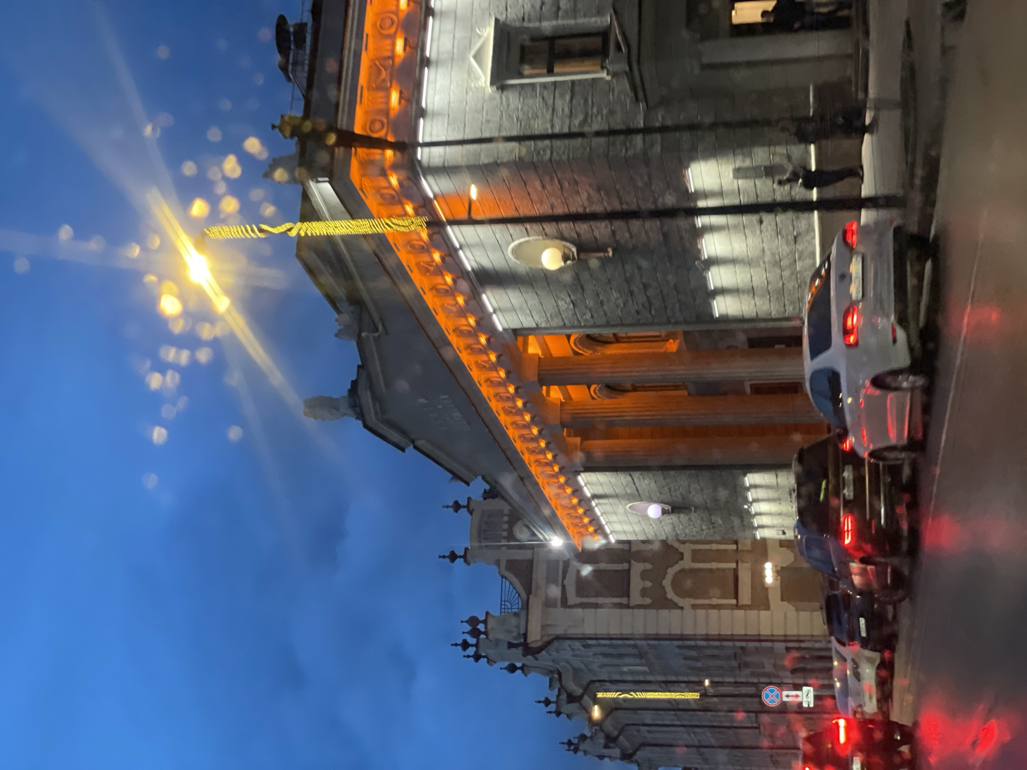 Here's a description of the image:
The image shows a street scene at dusk, likely in Krasnoyarsk, Russia based on building architecture. The perspective is from a low angle looking up at the buildings and street lights.
**Environment:** The sky is a deep blue, transitioning to lighter blue with some clouds. The streets are wet, reflecting light.
**Buildings:** Ornate architecture with decorative stonework is prevalent. There are multiple buildings visible, including a larger building with orange under-lighting that accentuates its architectural details (likely Krasnoyarsk State Opera and Ballet Theatre). Street lights are illuminated, and decorative lighting is present on at least one of the buildings.
**Objects:** Cars are parked in a row on the right side of the street, with their taillights illuminated. A stop sign can be seen. There are also street lights present and signs and decorations.
**People:** Two people are present in the image standing on the sidewalk.