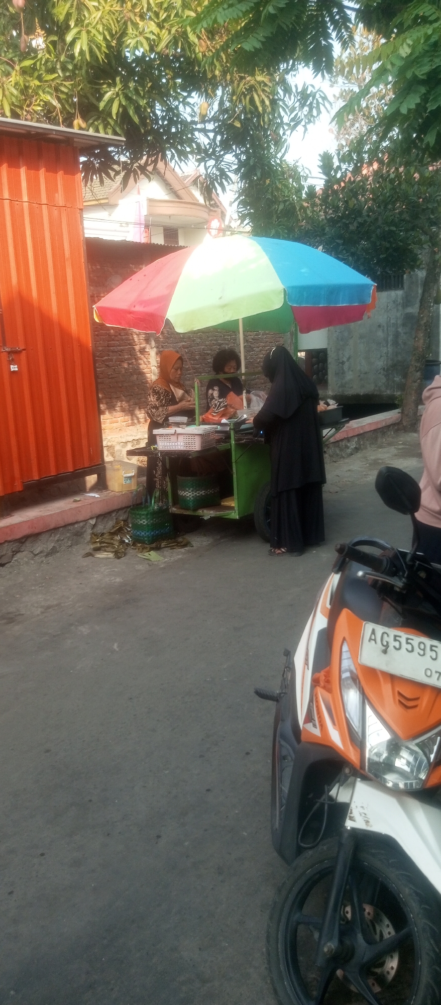 A street vendor is seen tending to a cart under a multi-colored umbrella. The vendor, wearing an orange headscarf and patterned shirt, is interacting with another individual partially obscured by the cart and wearing a black hijab and dress. A third person, wearing a black and white shirt, is also behind the cart. The cart is green and holds various items, including baskets and what appear to be food containers. In front of the cart, there are several green baskets and some food items laid out on the ground. To the left of the cart is a tall, orange, corrugated metal structure. The scene is set on a paved street, with a motorcycle in the foreground. The background shows brick walls and foliage. The license plate on the motorcycle reads "AG 5595 OS". The overall impression is of a busy street-side market. This scene is typical of street vending in Kediri City, Indonesia, where vendors often operate from carts and display their wares under umbrellas.