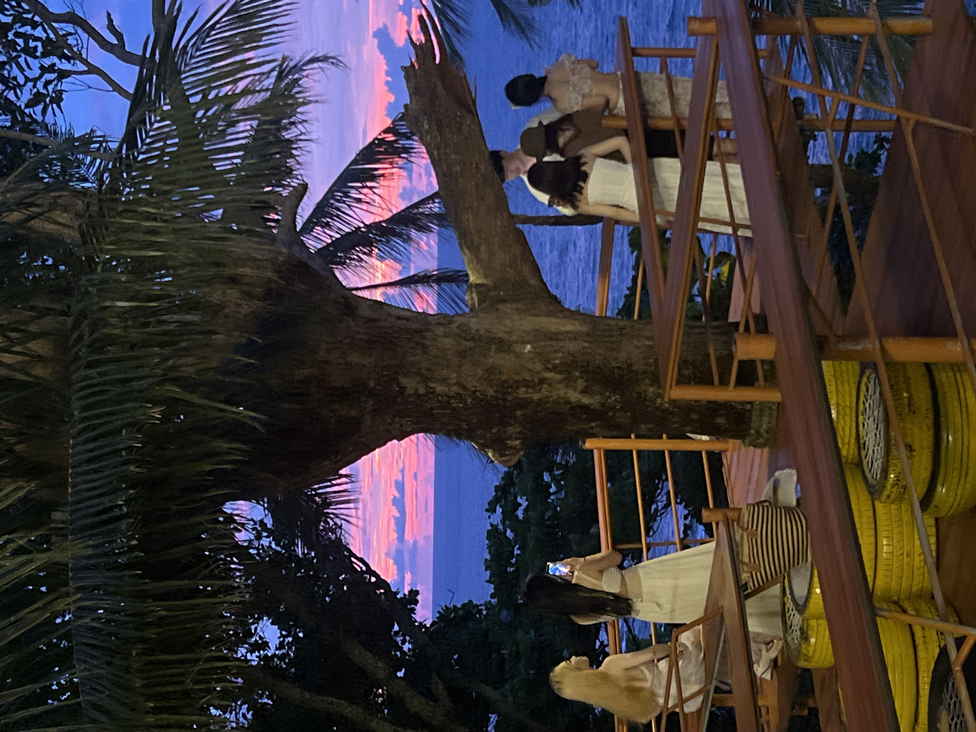 A group of young women are gathered on a multi-level wooden structure overlooking the ocean at dusk. The sky is a dramatic mix of deep blue and vibrant pink and orange, suggesting a beautiful sunset. Palm trees frame the scene, their fronds casting shadows. One woman in the foreground is taking a photo with her phone, while others are socializing and posing on the elevated platform. The structure appears to be part of an outdoor cafe or seating area, with visible railings and seating areas. Yellow tires are stacked in the background, adding an unusual decorative element. The overall mood is relaxed and convivial, with the women enjoying the scenic view and each other's company. The time of day is clearly evening, with the fading light casting a warm glow on the scene. The location cues, such as the palm trees and the ocean view, point to a tropical setting.