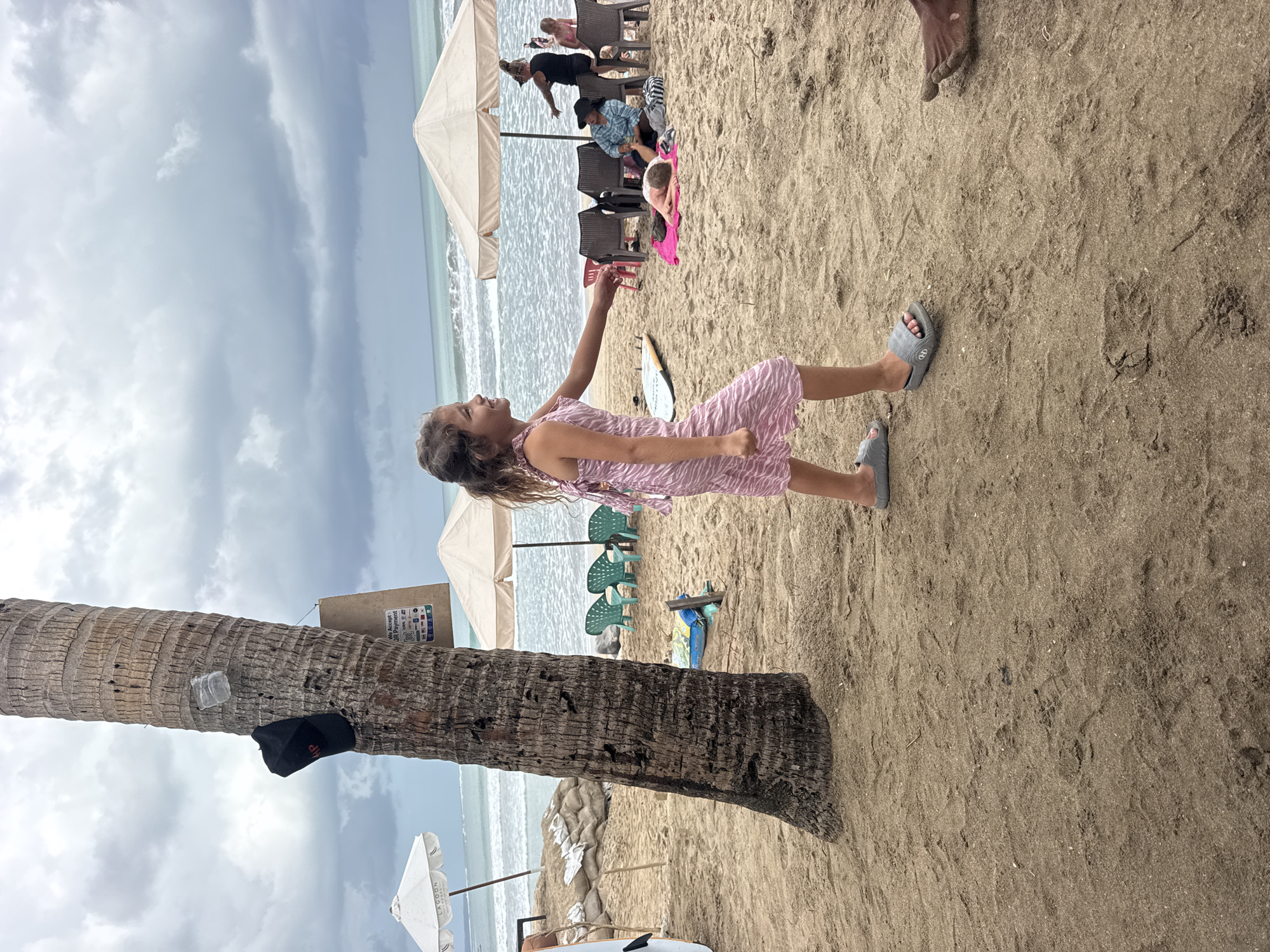 An outdoor, medium shot captures a young girl with curly brown hair wearing a pink and white striped dress and gray slides standing on a sandy beach in Kuta, Indonesia. She is positioned in the foreground, facing right, with her right arm extended. Behind her, a large palm tree trunk is visible with a black baseball cap and a plastic bottle attached to it. Further in the background, several beach umbrellas, chairs, and people are scattered across the sand, with the ocean and a cloudy sky completing the scene. The sand is textured with footprints.
