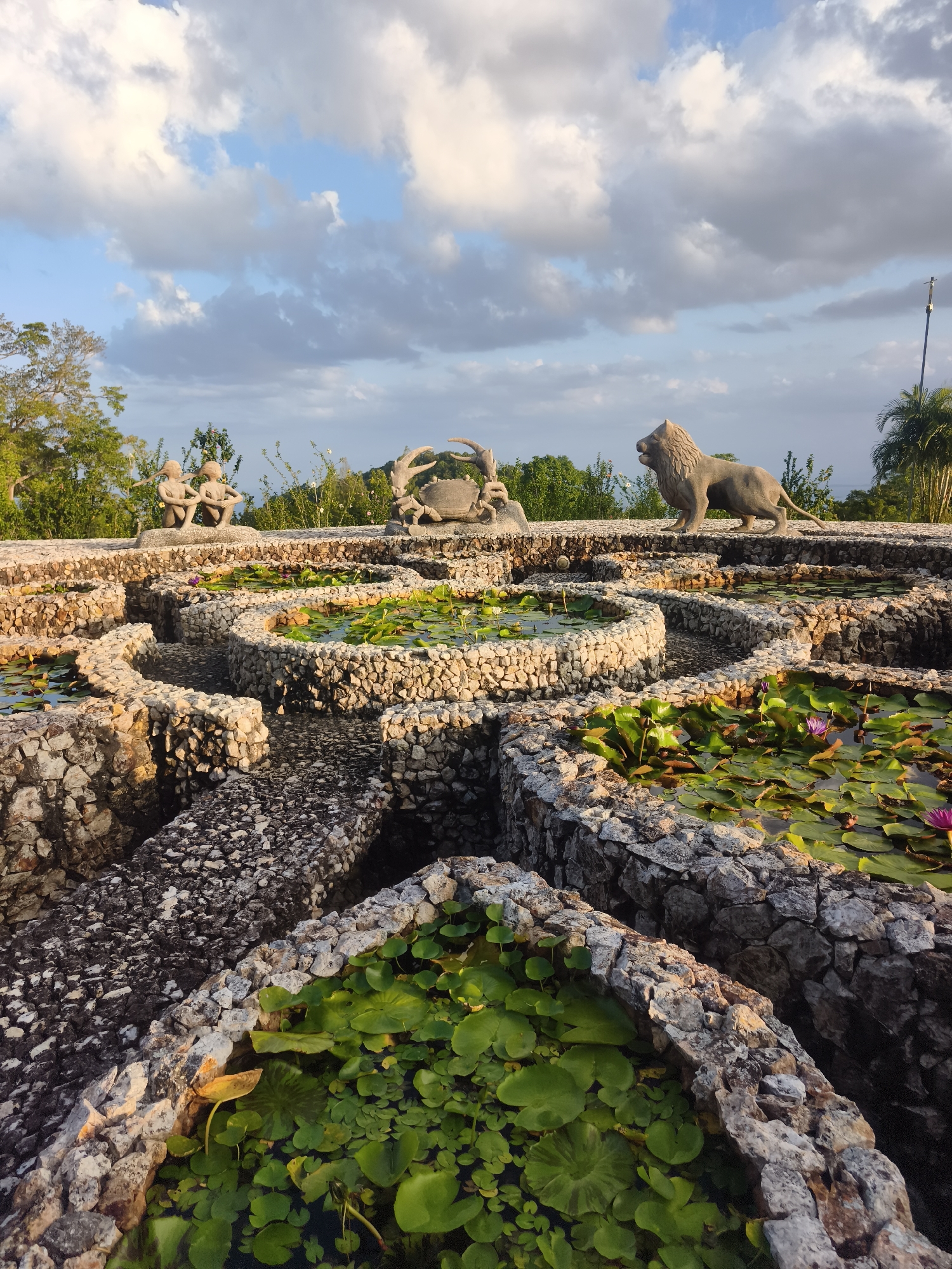 The image depicts a water maze garden located in Koh Samui, Thailand. The garden features multiple circular pools filled with water lilies and surrounded by stone walls made of rough-hewn rocks. Statues of various animals and figures are strategically placed throughout the garden. In the background, a statue of a lion stands prominently on a raised platform. To its left, a statue of a crab is visible, and further to the left, two seated figures resembling children can be seen. The environment is lush with green trees and foliage surrounding the garden. The sky above is filled with white and gray clouds against a blue backdrop, suggesting it is daytime.