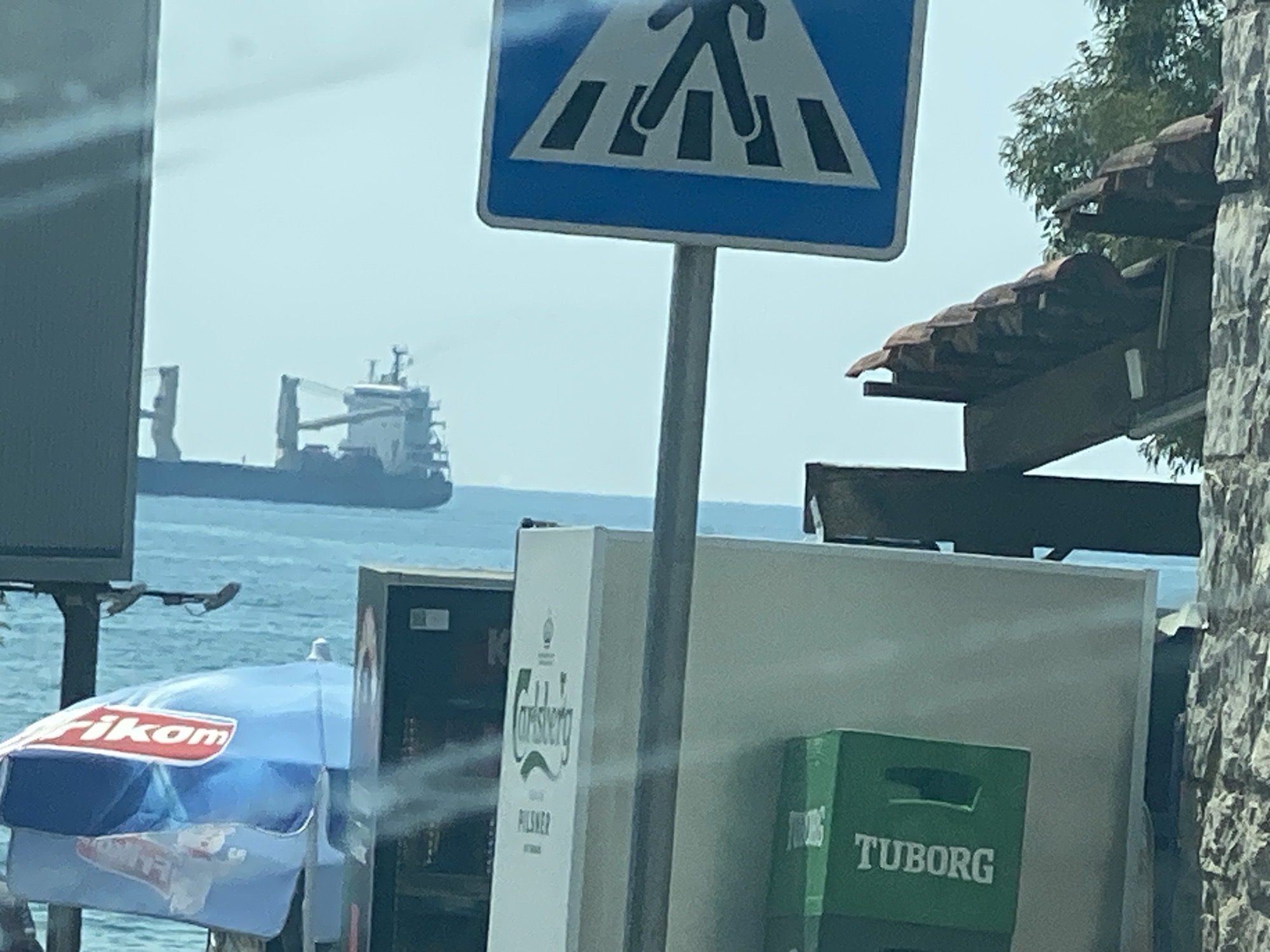 The image depicts a scene likely in Bar, Montenegro, featuring a pedestrian crossing sign prominently displayed in the foreground. In the background, a large cargo ship is visible on the calm, blue sea. Several objects are present in the middle ground, including a blue and white umbrella with red lettering that reads "Fikom," a white structure with "Carlsberg" and "Pilsner" branding, and a green crate with "Tuborg" branding. The environment suggests an outdoor setting, possibly near a waterfront area or a street with views of the sea. The lighting indicates it is daytime, with a bright, overcast sky. No people are clearly visible in the image, and no specific actions or interactions are depicted. The overall impression is a still, somewhat cluttered, outdoor scene.