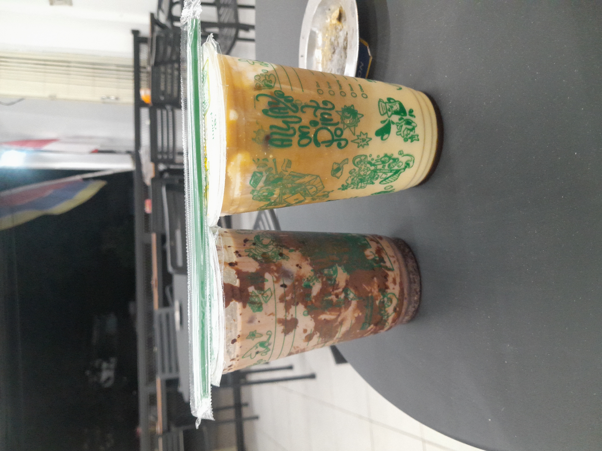 Two clear plastic cups filled with iced beverages are placed on a dark grey circular table. The cup on the left contains a chocolate-colored drink with swirls of lighter brown. The cup on the right contains a lighter brown beverage with a foamy top. Both cups have green plastic lids sealed with a clear plastic film, and a green plastic straw is placed horizontally across the top of each cup, resting on the film.
The cups are decorated with green patterns and text. The cup on the left has a pattern of what appear to be leaves and possibly small animals. The cup on the right has the text "ALWAYS ON POINT" in a stylized font, along with various green illustrations, including small figures and possibly cartoonish characters.
In the background, the scene appears to be indoors, possibly a cafe or restaurant. Dark-colored tables and chairs are visible, as well as a blurred section of a wall with what looks like a flag or banner hanging. The lighting suggests it is either evening or an indoor setting.
The image is taken from a slightly elevated angle, looking down at the table and the drinks. The focus is on the drinks, with the background being out of focus. There are no people clearly visible in the image. The scene is clean and orderly. The setting appears to be a public establishment, likely in Desa Ponokawan, Indonesia, given the context.
