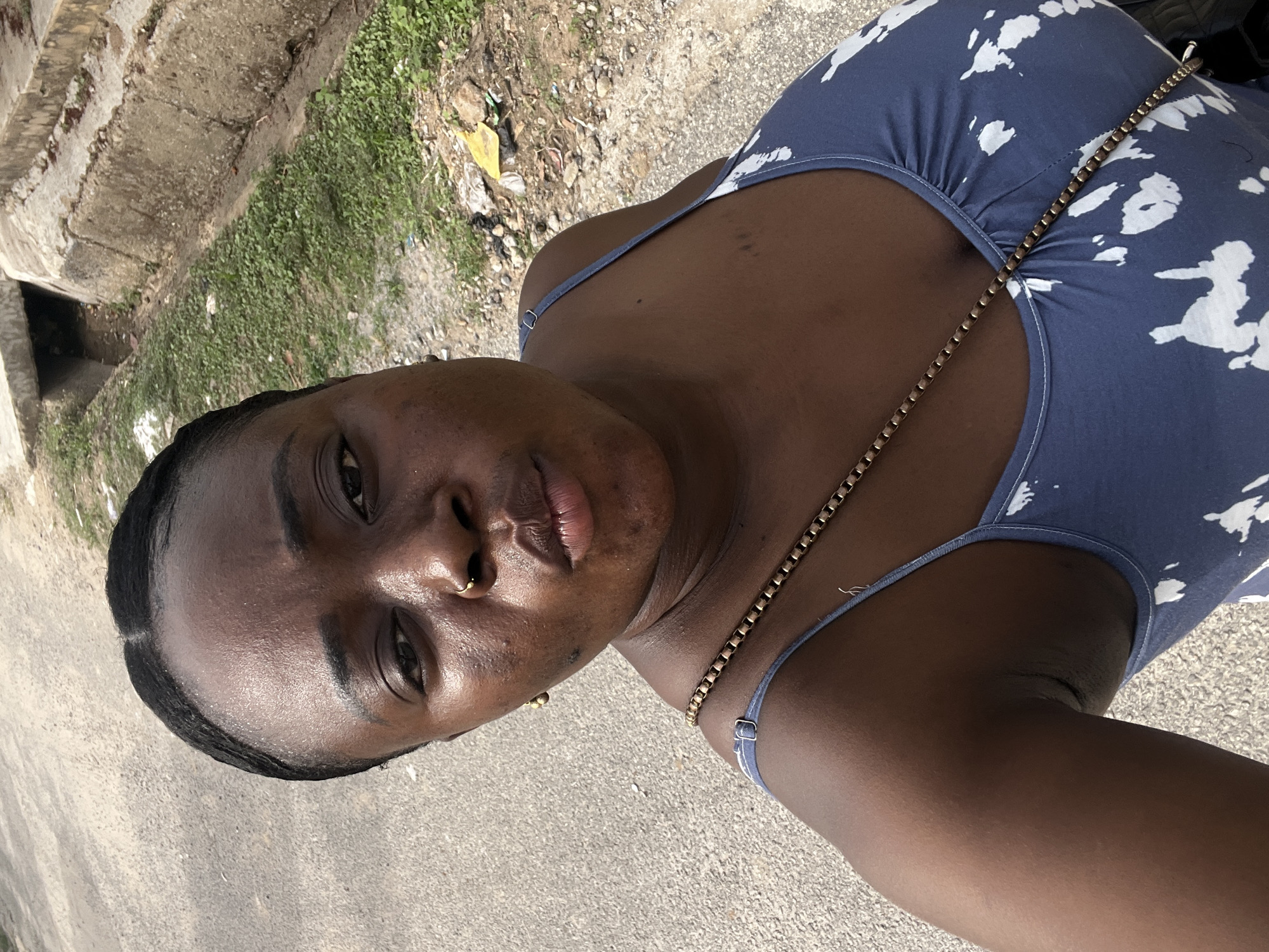 A young dark-skinned woman takes a selfie outdoors. She wears a blue patterned tank top and a gold chain. She has a nose ring in her left nostril and a gold earring in her left ear. The background is a concrete street and adjacent curb and vegetation. It is not possible to confirm the location as Abuja, Nigeria from the given visual information.