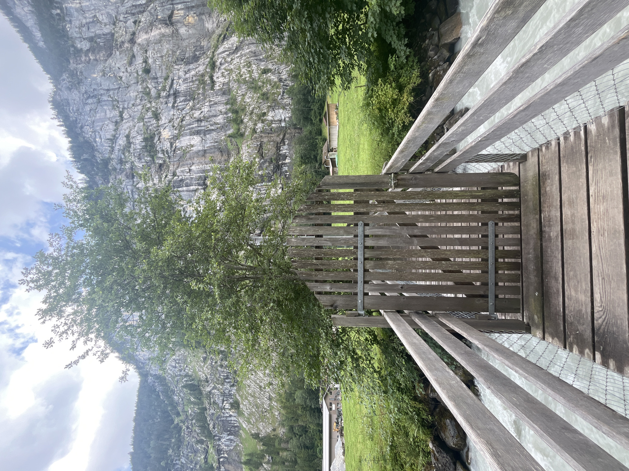 The image depicts a wooden bridge with a closed gate in the foreground, spanning a fast-flowing, milky-colored river. The bridge has wooden railings and mesh wire on the sides. Beyond the bridge, a lush green meadow is visible, dotted with trees and a few buildings. In the background, a towering, sheer rock face dominates the landscape, with a dense forest at its base. The sky is partly cloudy with patches of blue. The scene is set in Wengen, Switzerland. No people are visible in the image.