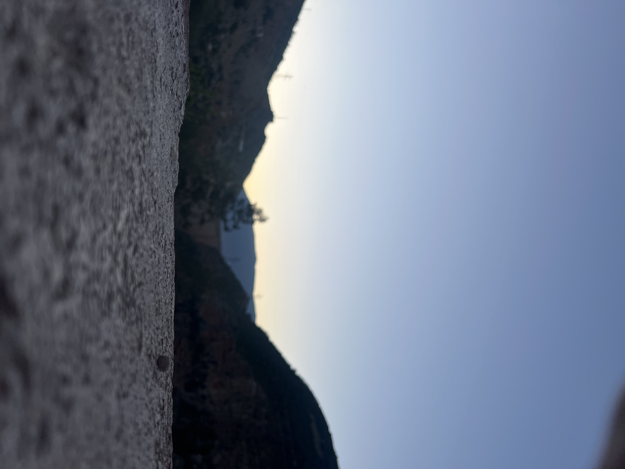 The image shows a view in Bin El Ouidane, Morocco, with a roughly textured wall in the foreground on the left. To the right, the silhouette of mountains and cliffs is visible against a light blue sky. A small tree or bush is nestled between the mountains. The angle suggests the photo was taken looking upwards.