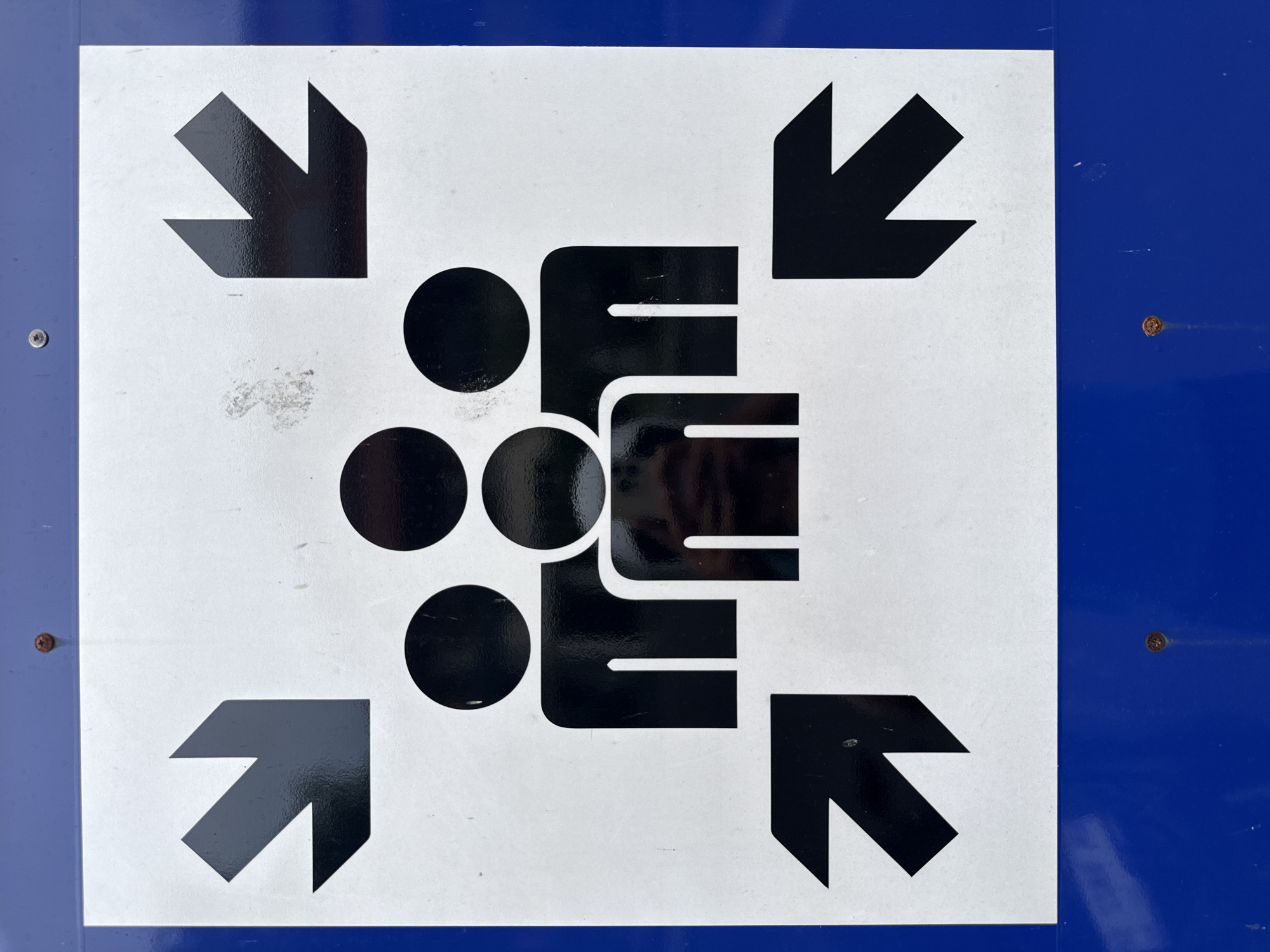 The image depicts a sign indicating a designated meeting point or assembly area. The sign consists of a white square with black graphics, mounted on a blue background. The graphic includes four arrows pointing inward from the corners of the square. In the center, there is a stylized figure of a group of people depicted as three circles stacked vertically next to a square shape with three horizontal lines, representing individuals. The sign is secured with four fasteners, and the surface shows signs of wear and slight discoloration. The location of this sign is Nusa Dua, Indonesia.