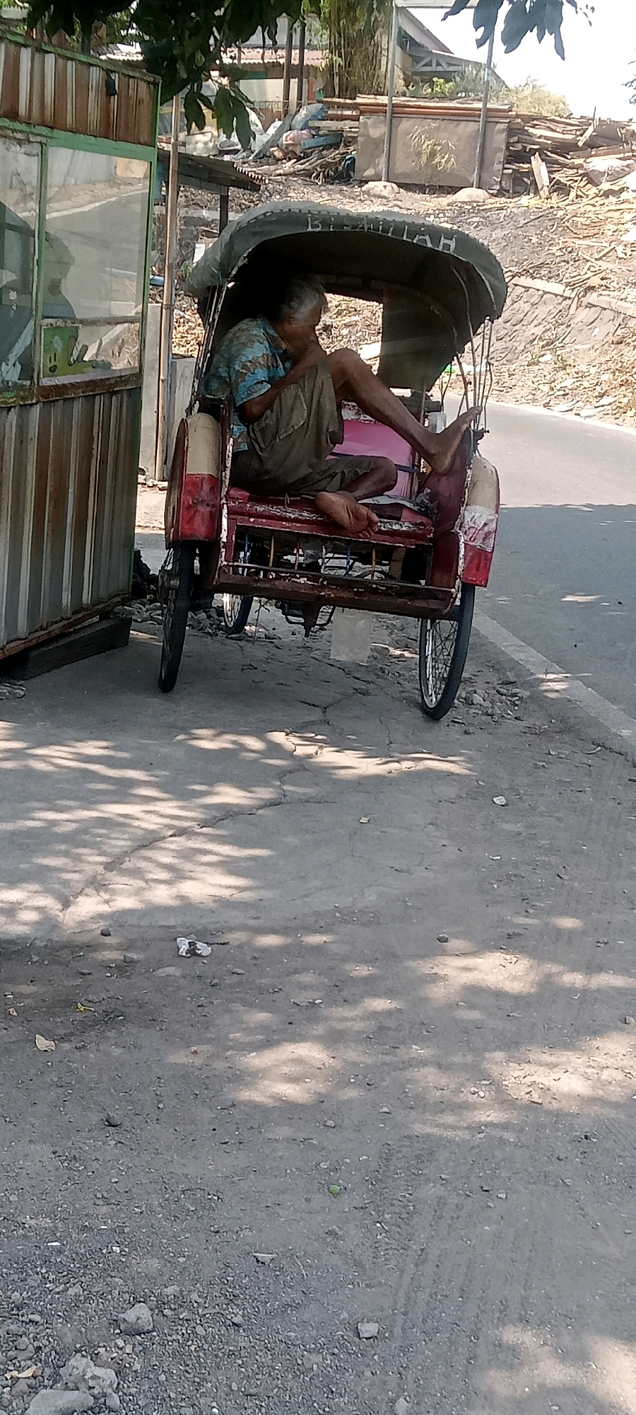 An elderly person is sitting inside a red and white auto-rickshaw parked on a cracked asphalt road. The auto-rickshaw has a green canopy with Arabic script written on it. The person is wearing a blue and white striped shirt and beige shorts, with their legs outstretched. The background shows a pile of debris and some vegetation. To the left of the auto-rickshaw is a rusty, corrugated metal structure with glass panels. The scene appears to be in Surakarta, Indonesia, given the visual context of the auto-rickshaw and surrounding environment.