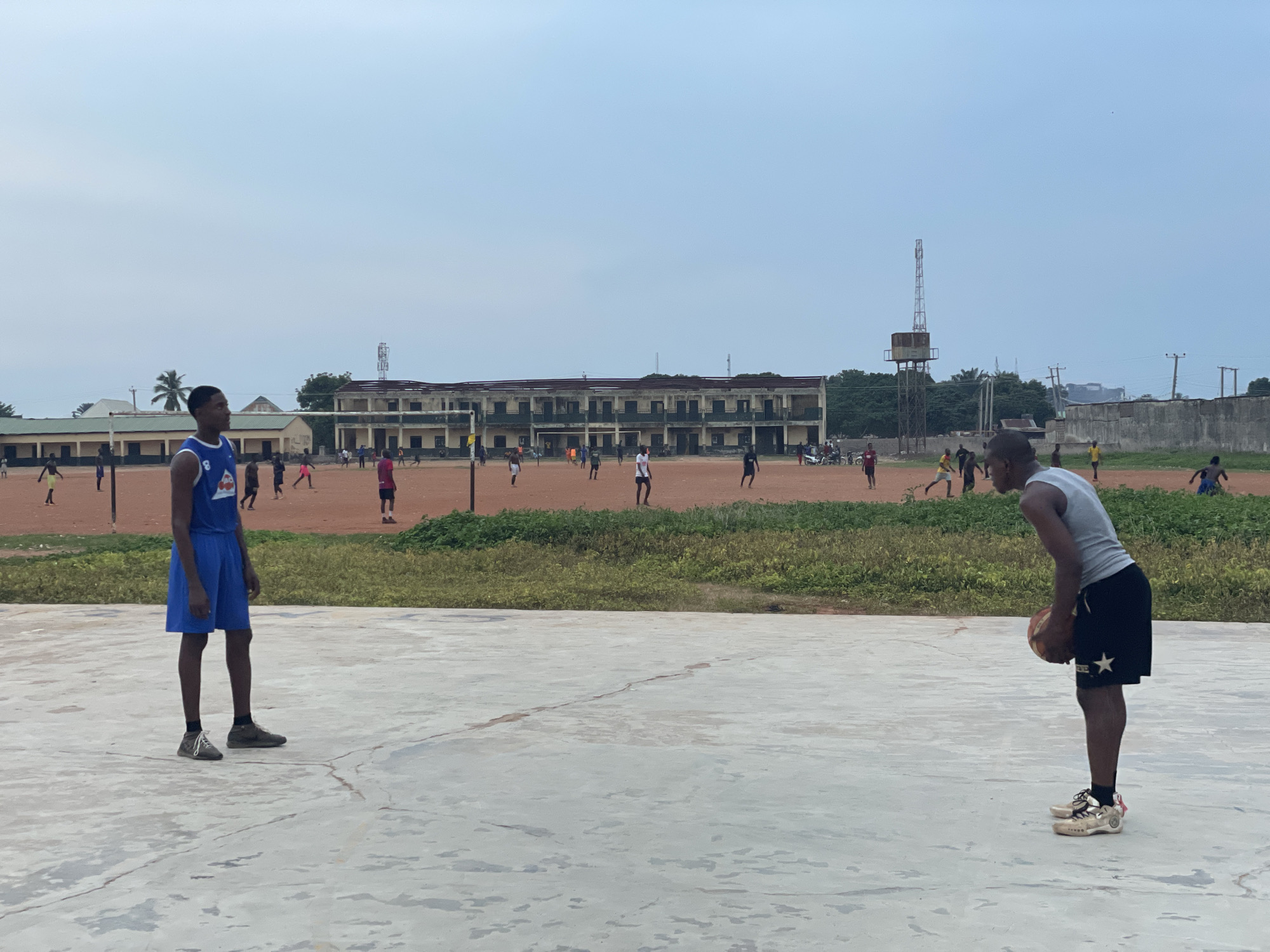 In Akwanga, Nigeria, a basketball court is the setting for an outdoor activity on an overcast day. Two young men are the primary subjects. One, wearing a blue basketball jersey with white trim and blue shorts, stands on the left side of the foreground, looking to the right. The other, in a gray tank top and black shorts with a white star emblem, is on the right side of the foreground, bent over holding a basketball.
In the middle ground, a red dirt field is visible, where a group of people, likely young men and boys, are engaged in a soccer game. They are scattered across the field, some kicking the ball, others running. Behind the soccer field, a long, two-story building with numerous windows and balconies dominates the background. This building appears to be a school or administrative complex. A telecommunications tower with a platform structure stands to the right of the building. The environment is generally dusty and open, with some patches of green vegetation visible. The sky is a pale, hazy blue, suggesting a humid or cloudy atmosphere.