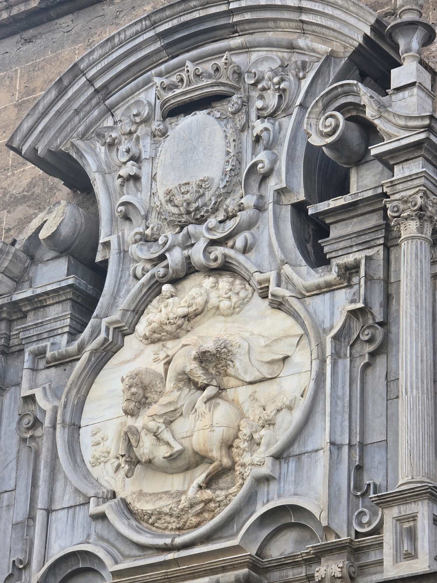 This is a low-angle, eye-level, outdoor, close-up, full shot of an ornate stone facade in Granada, Spain, likely a church or governmental building. The facade features a large, central relief sculpture framed by elaborate architectural elements. The sculpture depicts a winged figure, possibly an angel or mythological character, riding a horse. The figure is shown from the chest up, with flowing hair and drapery, and appears to be looking upwards. The horse is depicted in mid-stride, with its front legs raised. The background of the relief is a cloudy sky, with some smaller figures or cherubs above. The surrounding architectural elements include volutes, scrolls, acanthus leaves, and a large crest or coat of arms at the top. A fluted column with a Corinthian capital is visible on the right side of the image, and a rough stone wall forms the background on the upper left. The stonework shows signs of age and weathering, with some cracks and discoloration.