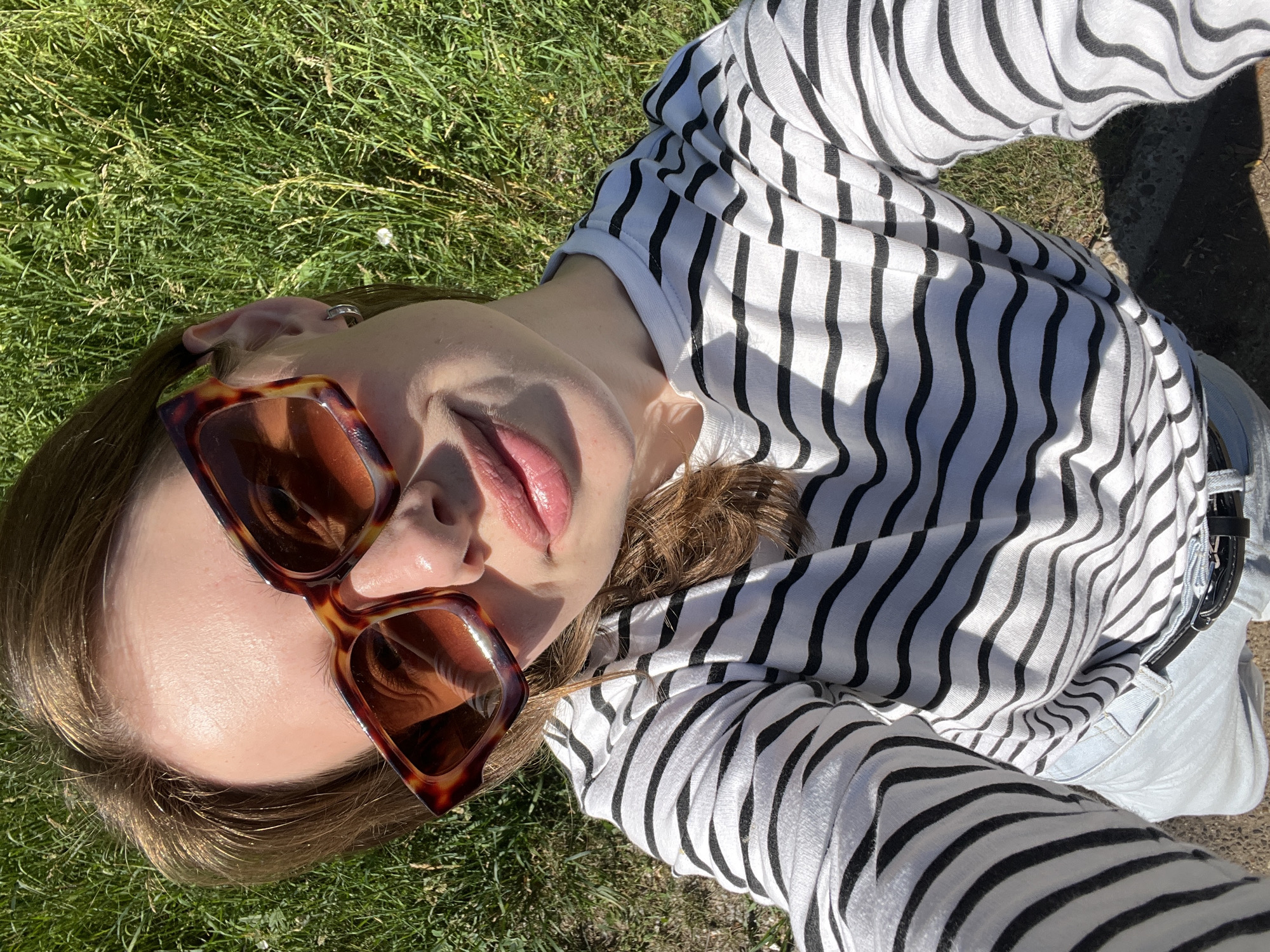 A young woman with light skin and brown hair is lying on grass, taking a selfie. She is wearing large, brown, square-shaped sunglasses with a tortoiseshell pattern, a white long-sleeved shirt with black horizontal stripes, and light blue jeans with a black belt. Her left ear is visible and she is wearing a small hoop earring. The background is lush green grass. The image appears to have been taken on a sunny day outdoors, likely in a park or a backyard. There is no discernible location information indicating Chaikovsky, Russia.