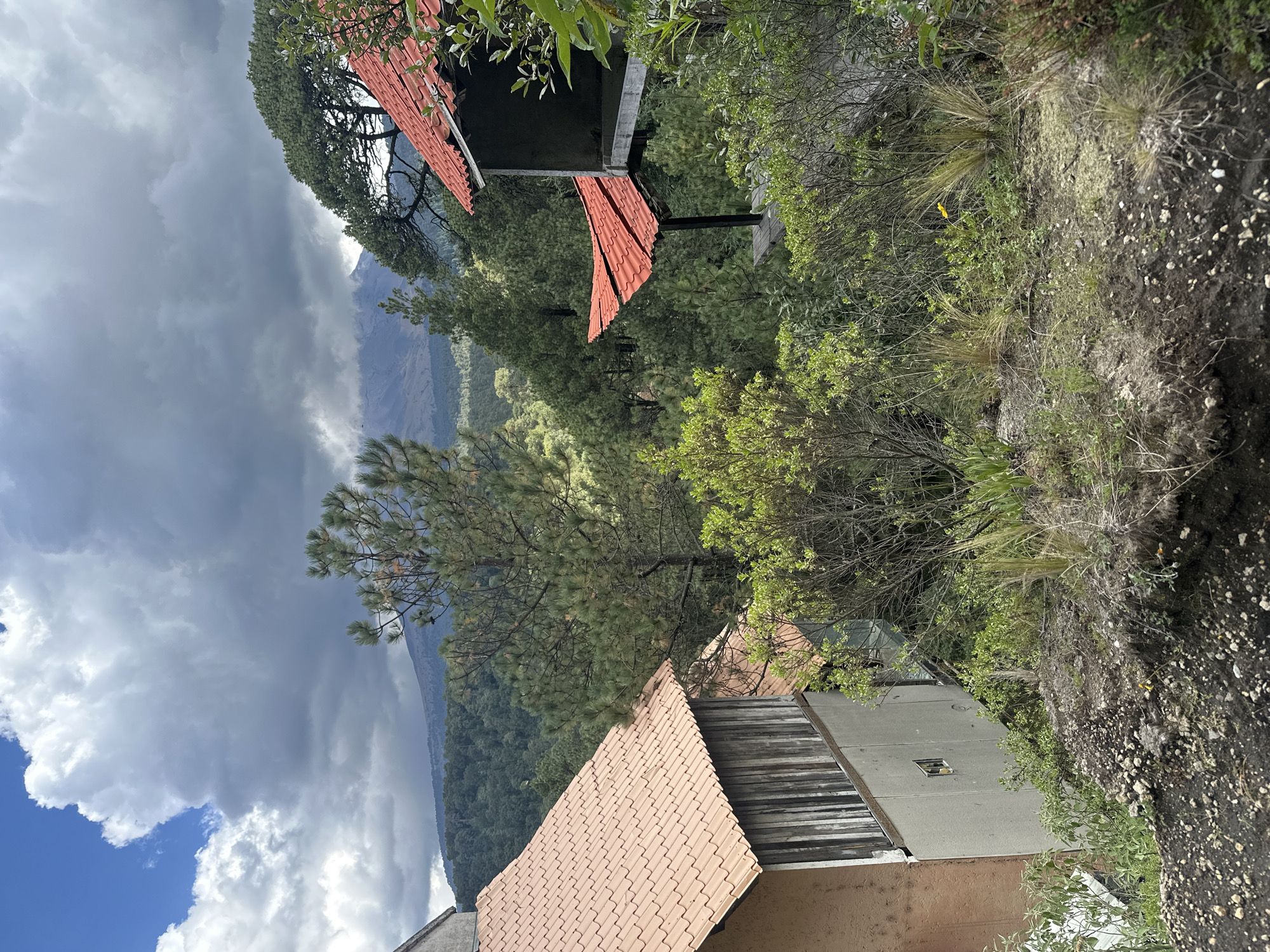 The image depicts a view from a hillside overlooking a forested mountain range with a volcano in the distance. In the foreground, there are two buildings with reddish-orange tiled roofs. The building on the left is partially visible and appears to be made of concrete and wood. The building on the right is a structure with an open side and a cantilevered roof, also with red tiles. The surrounding vegetation is lush and green, with various trees and bushes. The sky is partly cloudy, with a mix of blue sky and dark, dramatic clouds, some of which are obscuring the upper portion of the volcano. The volcano, identifiable as Popocatépetl, is visible in the background, with its peak shrouded in clouds and possibly volcanic smoke. The overall scene suggests a natural landscape in a somewhat remote or mountainous area, likely near Santiago Xalitzintla, Mexico, given the prominent view of Popocatépetl. There are no people visible in the image.