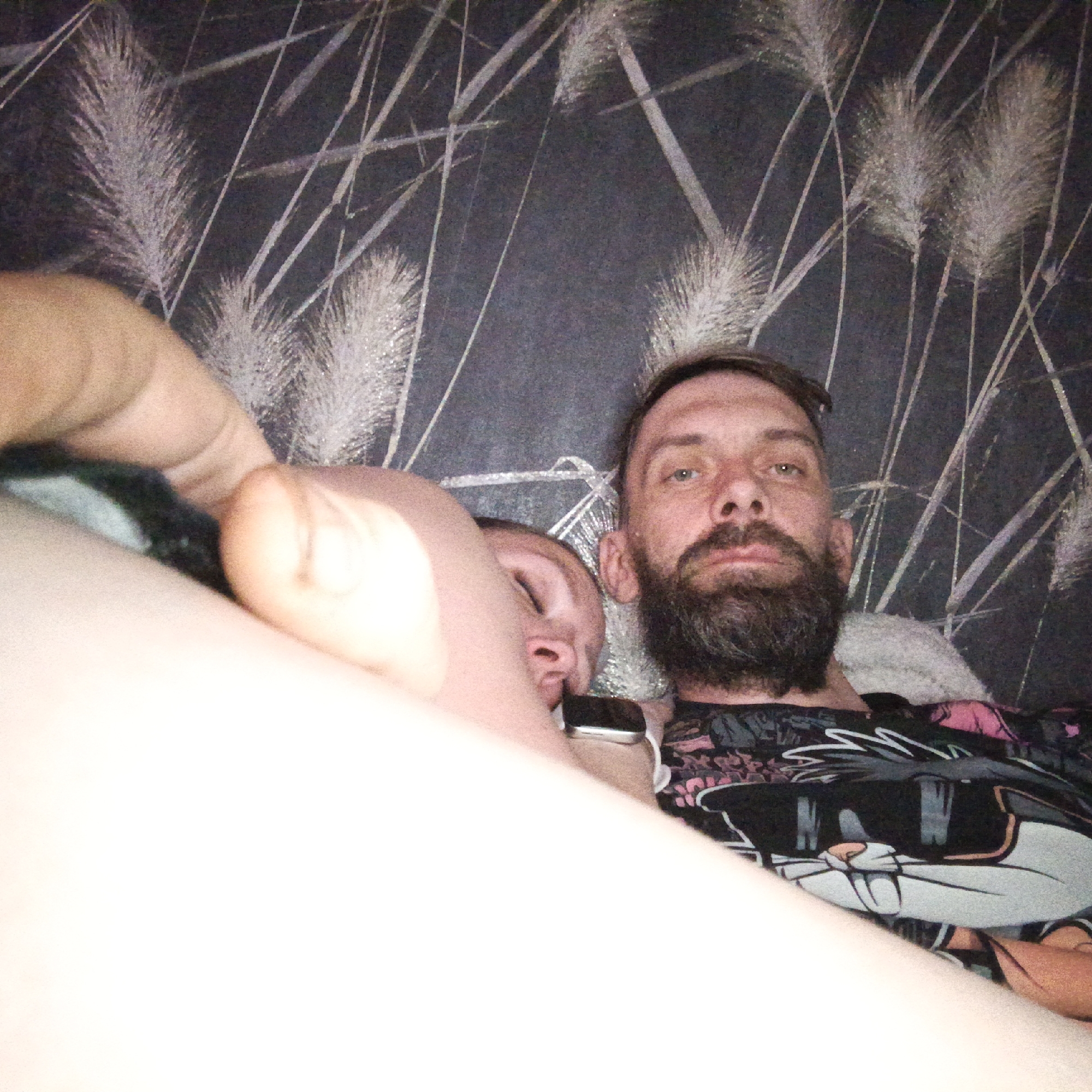 The image depicts two people lying down. A bearded man with fair skin faces the camera. Next to him, another person is lying down with their eyes closed. The man is wearing a dark shirt with a cartoon image. The background is a wall with a patterned wallpaper resembling wheat or grass stalks. The location is unclear. There is no indication of being in the city of Марковский, Russia.
