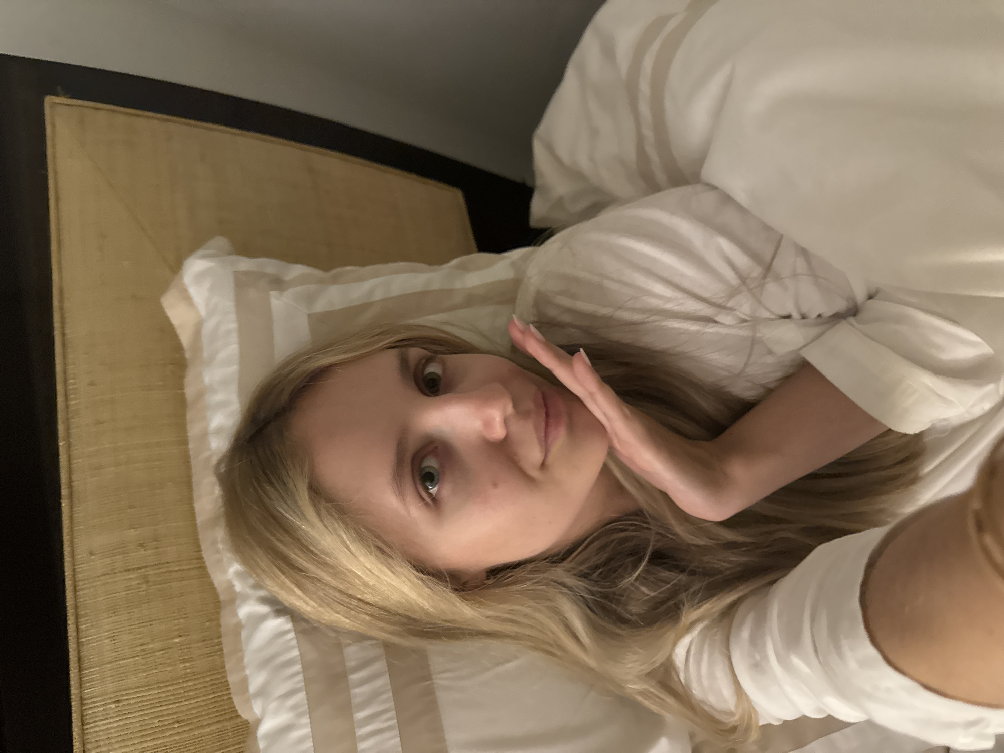 A young blonde woman lies in a bed, partially covered by white linens. She is wearing a white garment. Her right hand is raised, with fingers gently touching her chin. She is looking directly at the camera, and her expression is relaxed. The background shows a headboard made of a woven, light-colored material, and a dark wood frame. The overall lighting suggests an indoor setting, possibly a bedroom. The image does not contain any visual elements that would indicate a specific location in Ciudad de México, Mexico.