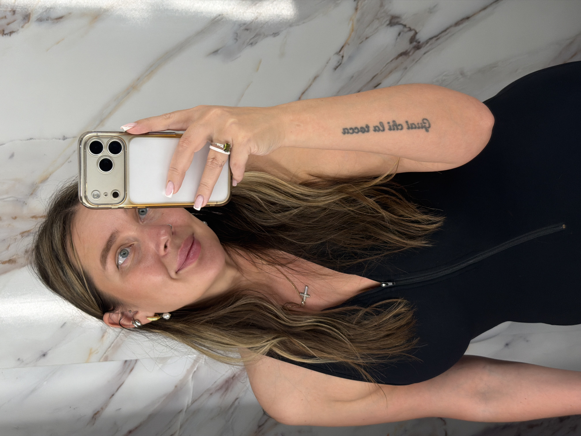 A person is taking a selfie. The person is a fair-skinned female with light brown hair, styled in waves. She is wearing a dark-colored, form-fitting top. She is holding a white and gold-colored iPhone in her right hand, using it to capture the image. A ring is visible on her right ring finger. Her left arm is partially visible, bearing a tattoo. She is wearing a necklace with a cross pendant, multiple earrings, and has manicured nails. The background is a marble-patterned surface, suggesting a wall or countertop. The location is not discernable as Moscow, Russia.