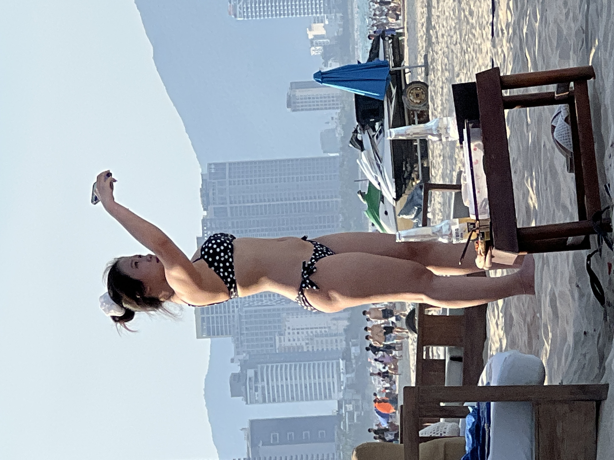The image shows a woman in a black and white polka dot bikini standing on a beach in Da Nang, Vietnam. She is holding a phone in her outstretched right hand, possibly taking a selfie or recording a video. Her left arm is bent, with her hand resting on her hip. In the background, there are tall buildings, mountains, and other beachgoers. There are also jet skis and a blue umbrella on the sand. A small table in the foreground holds two glass bottles and some food items. The lighting suggests it is daytime, likely late afternoon or early evening, given the soft light and long shadows. The overall scene depicts a casual day at the beach in a bustling coastal city.