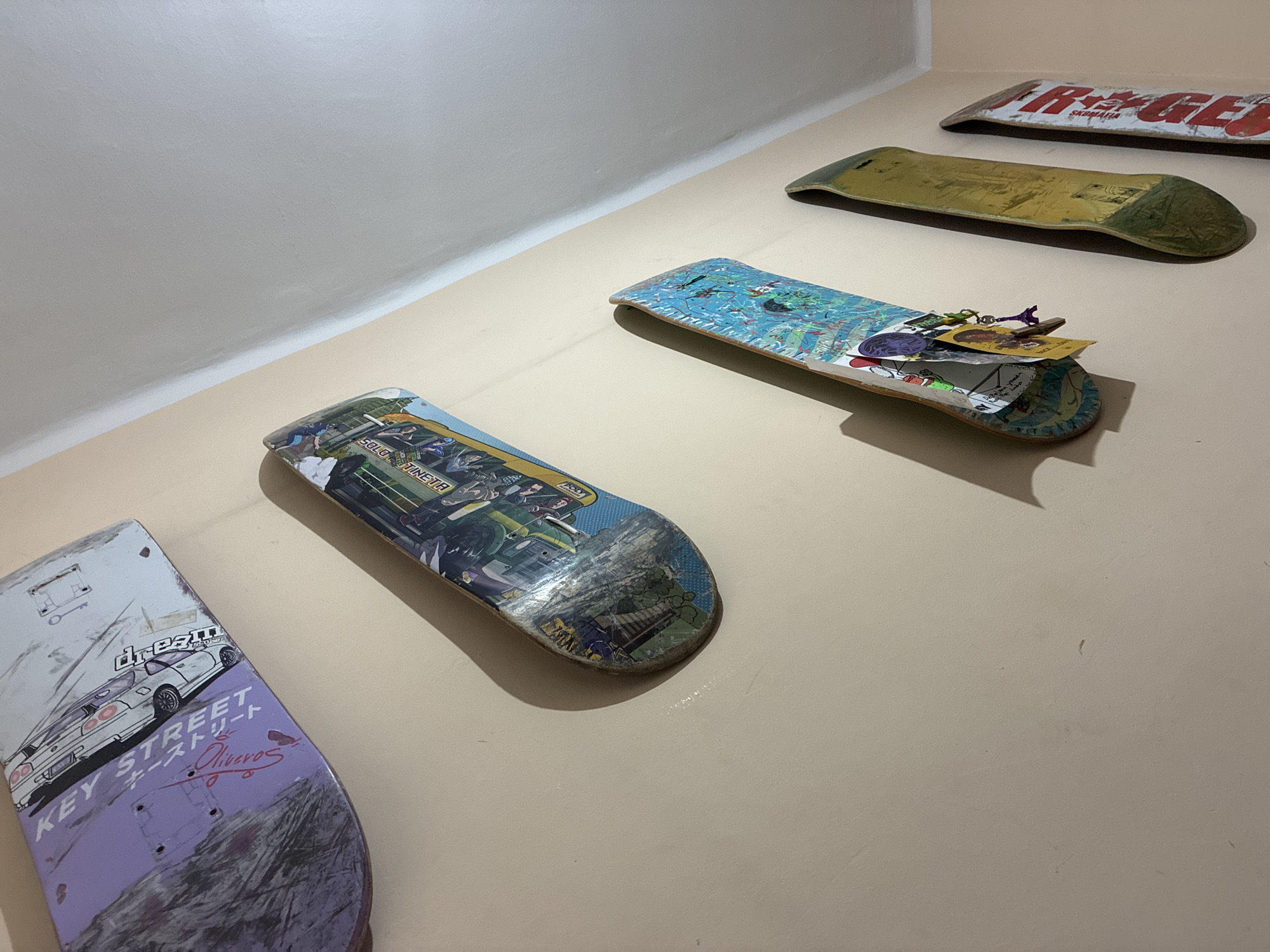 The image shows a collection of five skateboards mounted on a light-colored wall. The skateboards are arranged in a diagonal line from the top left to the bottom right of the frame. The topmost skateboard has a purple and white design featuring a white car and text that appears to read "KEY STREET". The next skateboard down has a colorful graphic with cartoonish figures and text that is partially obscured but includes "TINETR". The third skateboard is primarily blue with various abstract designs and some text. The fourth skateboard is yellow with green accents and appears to be older, with visible wear. The bottommost skateboard has red and white lettering. The wall appears to be interior, and the lighting suggests an indoor setting, possibly a room in l'Hospitalet de Llobregat, Spain, where the skateboards are displayed as art or decor. No people are visible in the image, and the skateboards are stationary.