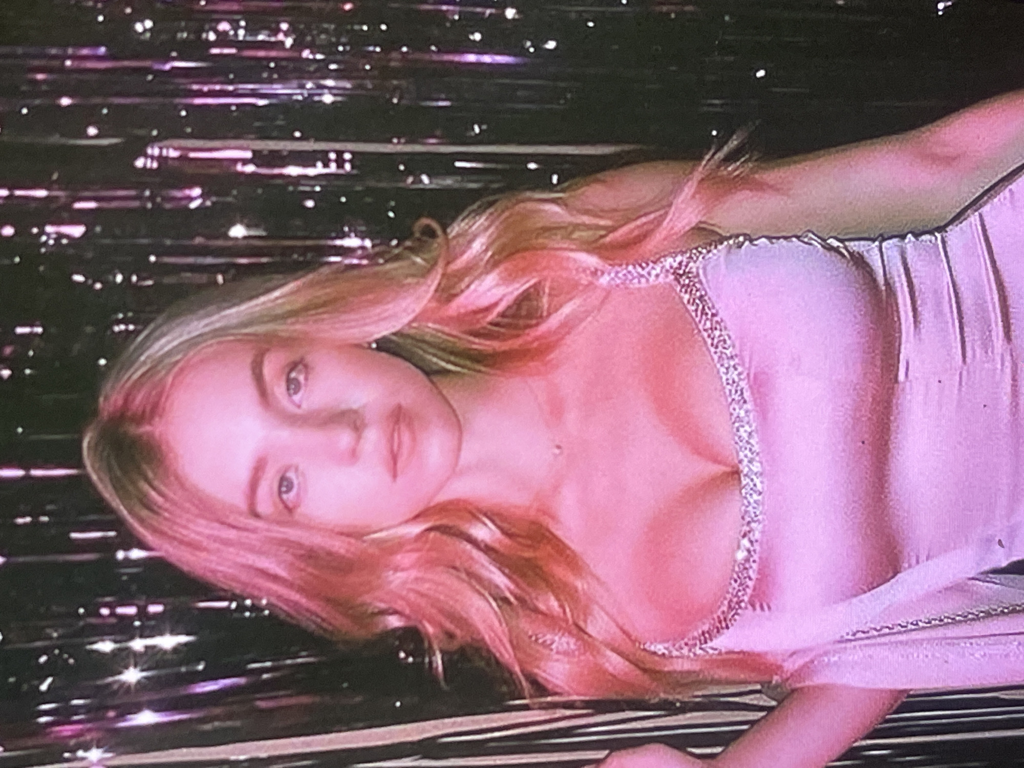 An overhead shot features a young woman with long, wavy blonde hair, which takes on a prominent pinkish hue from the ambient lighting. She is lying down, positioned horizontally across the frame, looking directly at the viewer with a calm or soft expression. She is wearing a light-colored (likely pink or pale purple) garment, possibly a dress or top, detailed with a sparkling or rhinestone-embellished strap or trim visible across her chest.
The background is dark and appears to be made of numerous horizontal, shimmering reflective strips, resembling tinsel or foil curtains, which scatter many small, bright, star-like light reflections. This creates a festive, sparkly, and somewhat dreamy atmosphere. The entire scene is bathed in a strong pink and purple light filter, giving the image a stylized and ethereal quality. The image's overall aesthetic suggests an indoor photoshoot or a party setting. There is no visible text. This scene is set in Poltava, Ukraine.