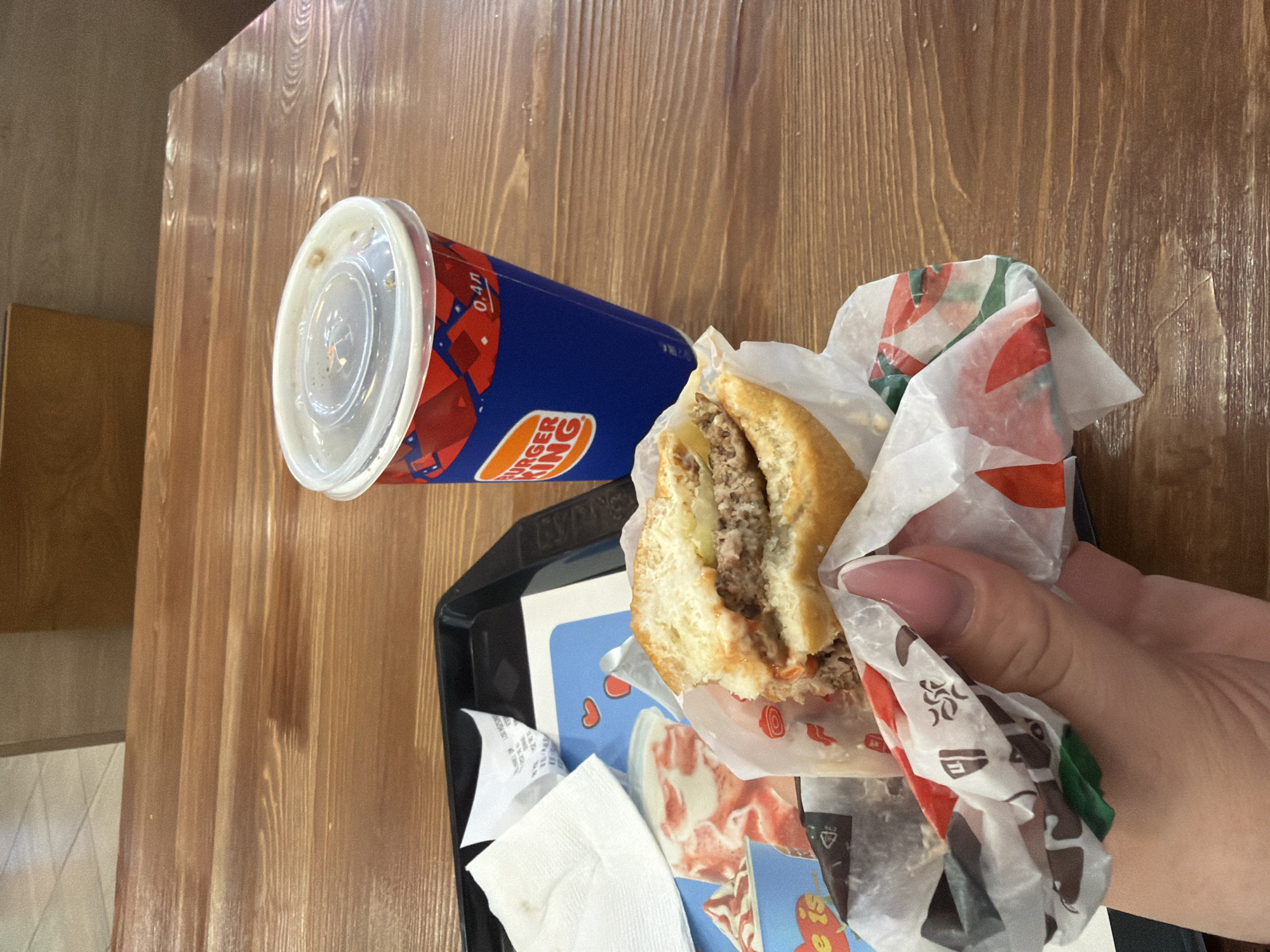 A Burger King meal, consisting of a partially eaten burger and a beverage, is presented on a wooden table. The burger, wrapped in branded paper, is held by a person's hand, revealing its meaty interior. Next to it, a blue Burger King cup, with its lid on, lies on its side. The cup features the Burger King logo and the text "0.4л". The meal is placed on a black tray, which also holds napkins and other paper items. The background shows a wooden tabletop with a textured grain. The scene appears to be indoors, possibly in a fast-food restaurant. The lighting suggests daytime.
