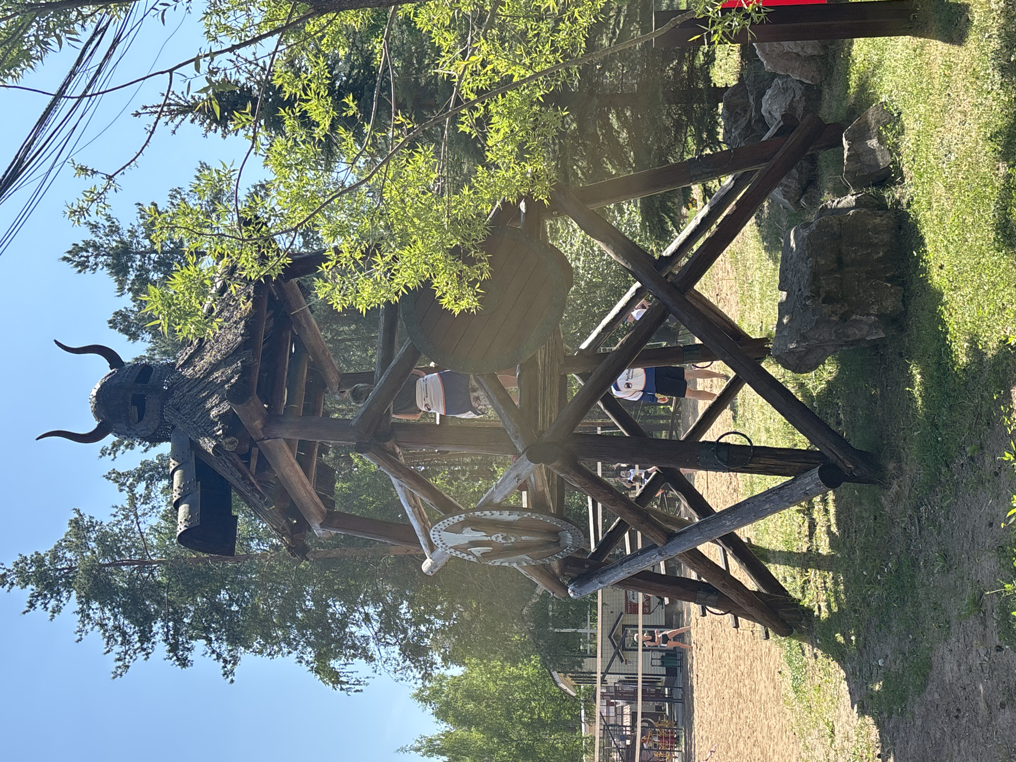 The image depicts a wooden play structure, designed in a Viking ship theme, located in the city of Chaykovsky, Russia. The structure includes a Viking helmet with horns mounted on top, a ship's wheel, and a large wooden barrel-like component. The structure is constructed of dark wooden beams and supports, arranged in a cross-braced pattern. People are visible inside the structure. The surrounding environment includes trees, grass, rocks, and sand. A volleyball court is visible in the background. The sky is blue.