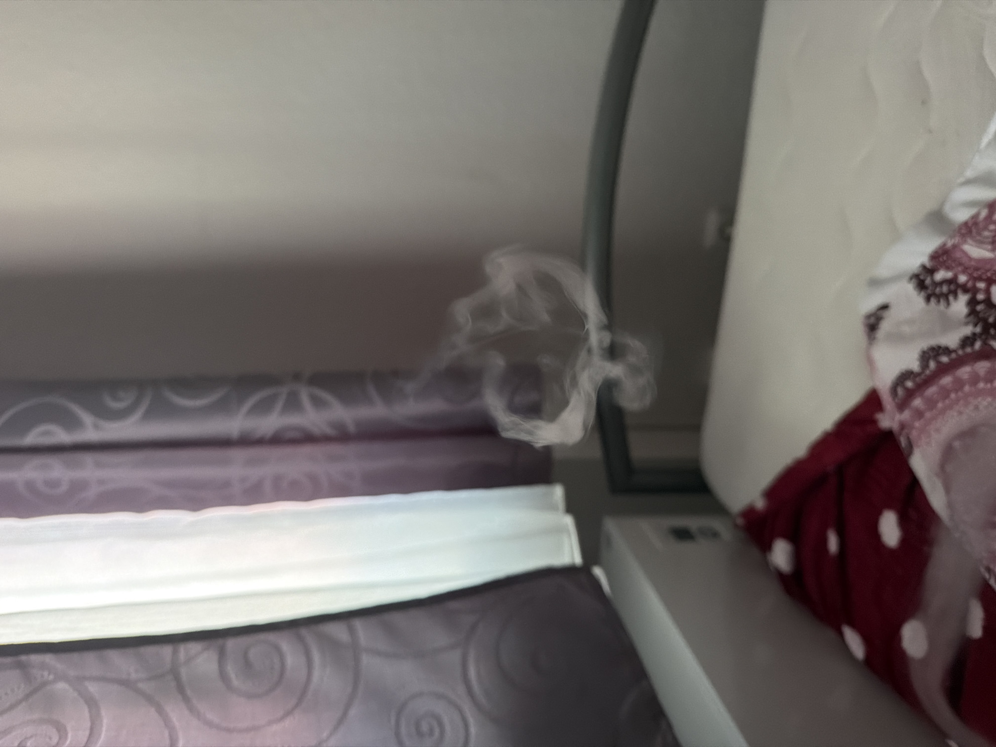 The image depicts a close-up view of a bed with layered bedding. The bedding appears to be in shades of purple and white, with patterned textures. A wisp of white smoke is visible in the upper center of the frame, swirling in the air. To the right of the bed, a white object, possibly a bedside table or piece of furniture, is partially visible, along with some fabric with a white polka dot pattern and decorative trim. The overall lighting suggests an indoor setting, potentially in a bedroom. The activity taking place is not explicitly clear due to the focus on the bedding and the presence of smoke, but it could imply someone has recently smoked or used an item that produced smoke in the room. No specific time of day or weather conditions can be inferred. There is no visible text in the image. The setting appears to be a domestic environment.