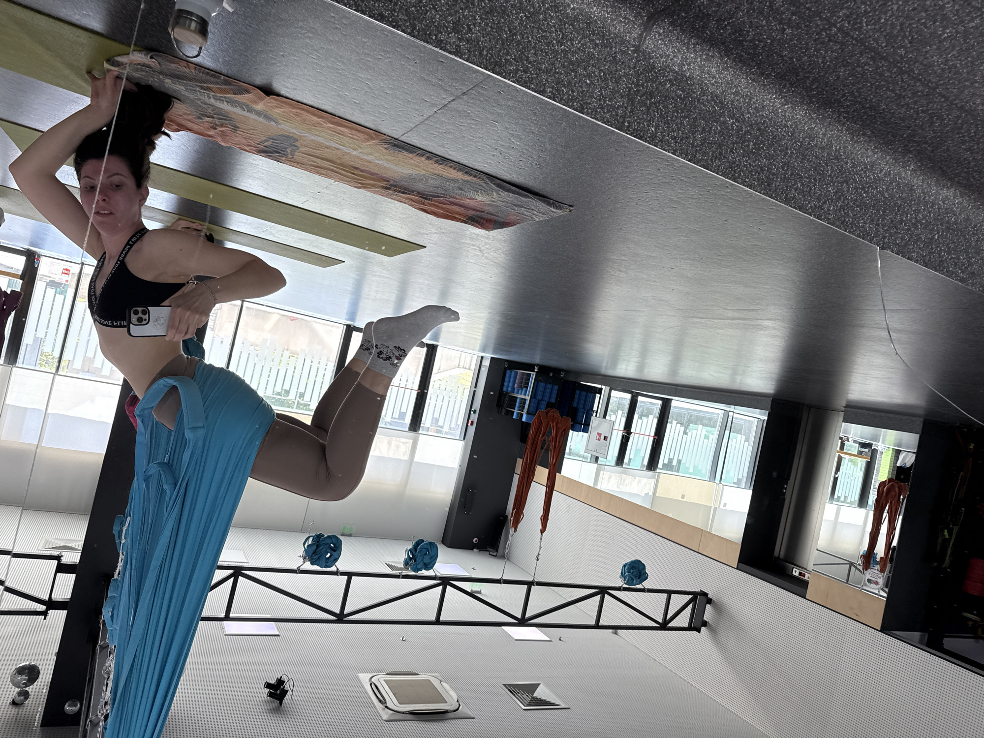 A woman is suspended in the air, seemingly practicing aerial yoga or a similar acrobatic activity, in a large, modern, and well-lit indoor space with high ceilings, the city is Unknown, Unknown. She is wearing a black sports bra and leggings, and she is holding a phone in one hand. She is secured in a blue fabric hammock or swing that hangs from the ceiling. The room features large windows, a black metal frame structure that appears to support hanging equipment or artwork, and a variety of objects on the floor, including what may be ventilation systems, equipment and storage, and possibly artwork. The floor is light-colored, and the ceiling is a mix of gray surfaces with textures or patterns. There's also a mat attached to the ceiling in the top center of the image.