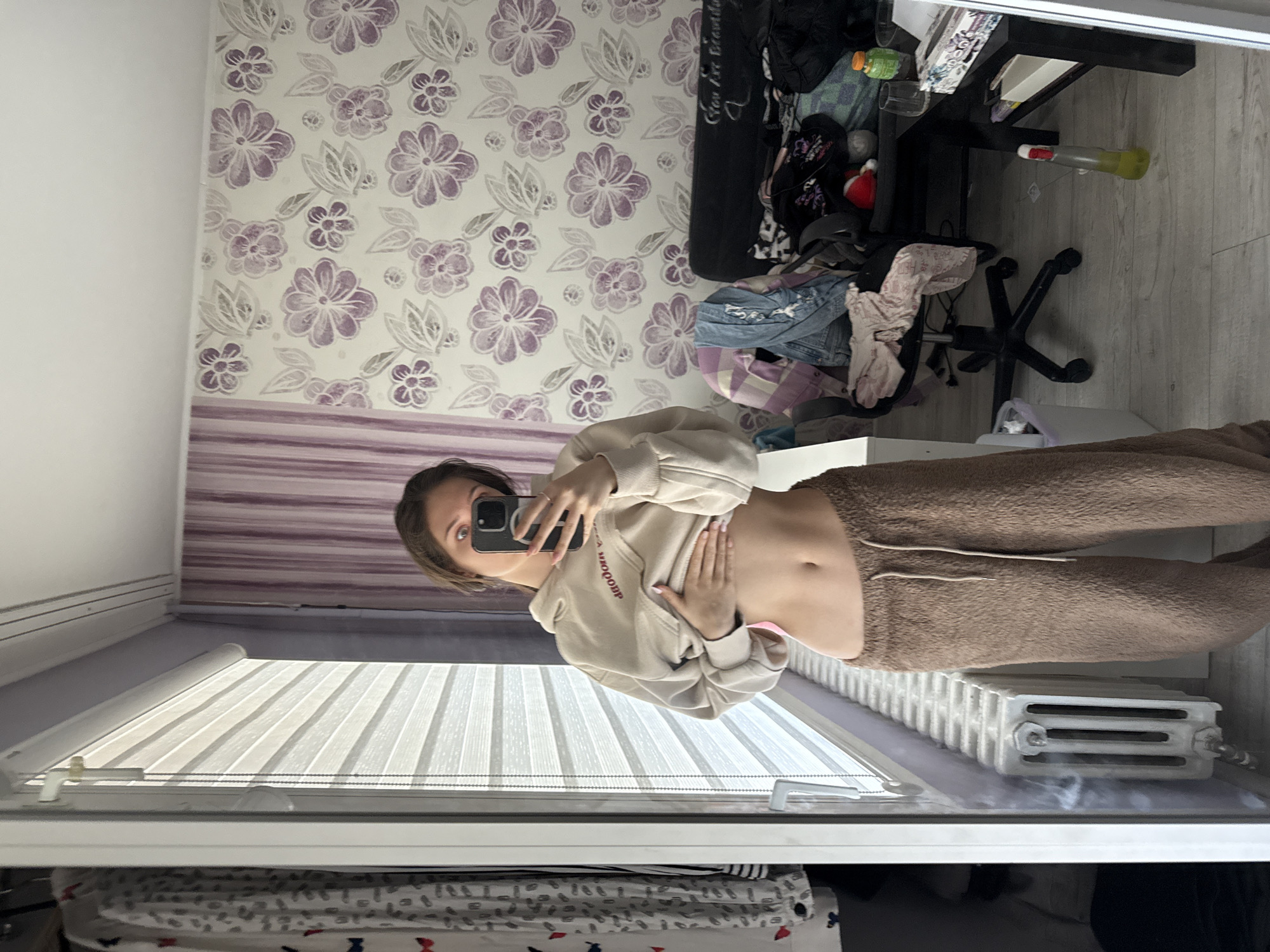 A woman is taking a selfie in a mirror. She is wearing a light beige hoodie and brown sweatpants, with her stomach exposed. The reflection shows a room with floral wallpaper, a desk cluttered with various items, and a radiator. Based on visual cues such as European-style blinds and a radiator, the location is likely in Europe. Without additional information, the specific city of Sofia, Bulgaria, cannot be confirmed.
