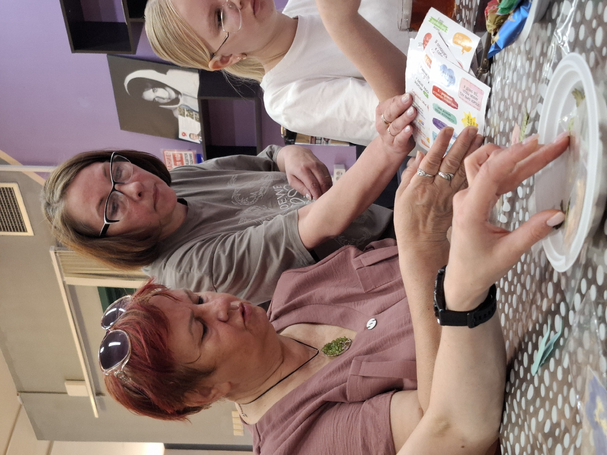 Three individuals are engaged in an activity at a table covered with a polka-dot tablecloth. A fair-skinned woman with glasses and light blonde hair is partially visible and appears to be examining a document. A woman with auburn hair and sunglasses pushed atop her head is in conversation, while another woman with reddish-brown hair, also wearing glasses, is observing the scene. On the table, there's a white plate, possibly containing items related to the activity. In the background, a purple wall with two black shelves and an unfocused black-and-white portrait can be seen. The location is likely in Markovskii, Russia, as the text in the background appears to be in Russian.
