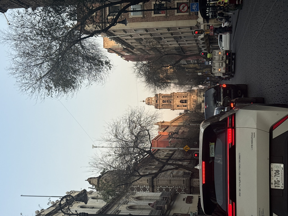A street view in what appears to be Mexico City, with a historical building, possibly a cathedral, visible in the distance. A white car with a visible license plate "17G-706" and "CDMX" (Ciudad de México/Mexico City) on the license plate, is stopped in traffic. Traffic lights are red. Trees with bare branches frame the view. Other vehicles, including a truck with workers, are also stopped in traffic. Street signs with bicycle symbols can be seen. Buildings line both sides of the street.