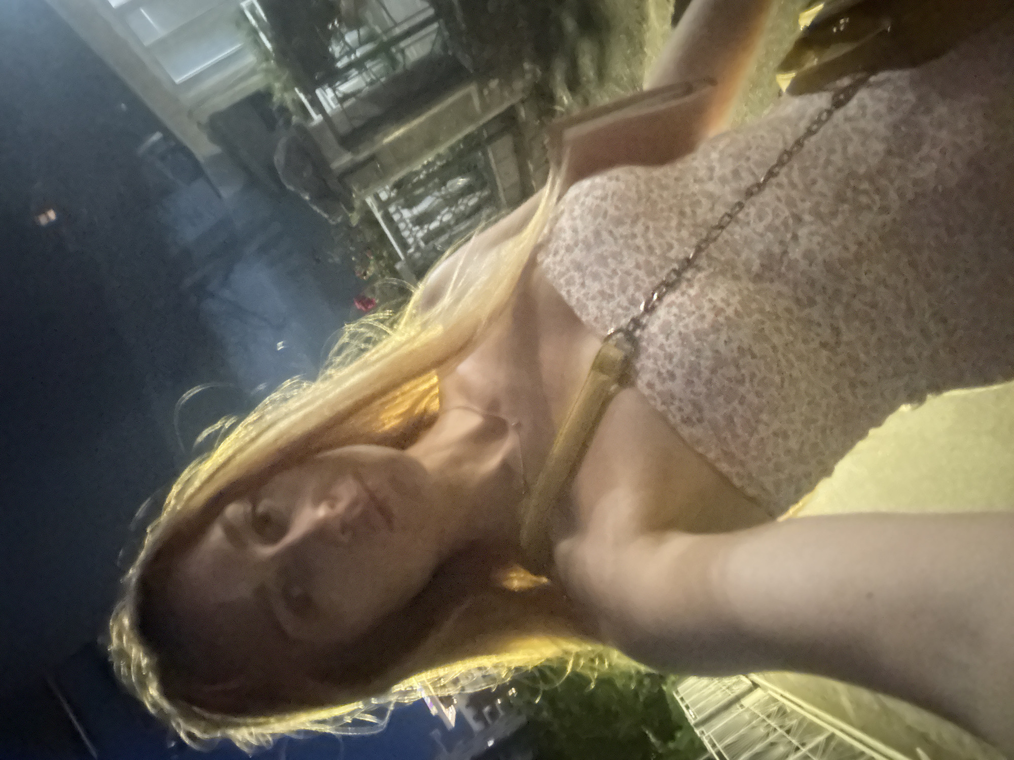 A young woman with long blonde hair is taking a selfie at night outdoors, likely in Budva, Montenegro. She is wearing a strapless dress with a floral pattern and a silver necklace. A yellow purse is slung across her body. The background appears to be a street with buildings and greenery. The lighting suggests it is nighttime, possibly with streetlights or ambient light from buildings.