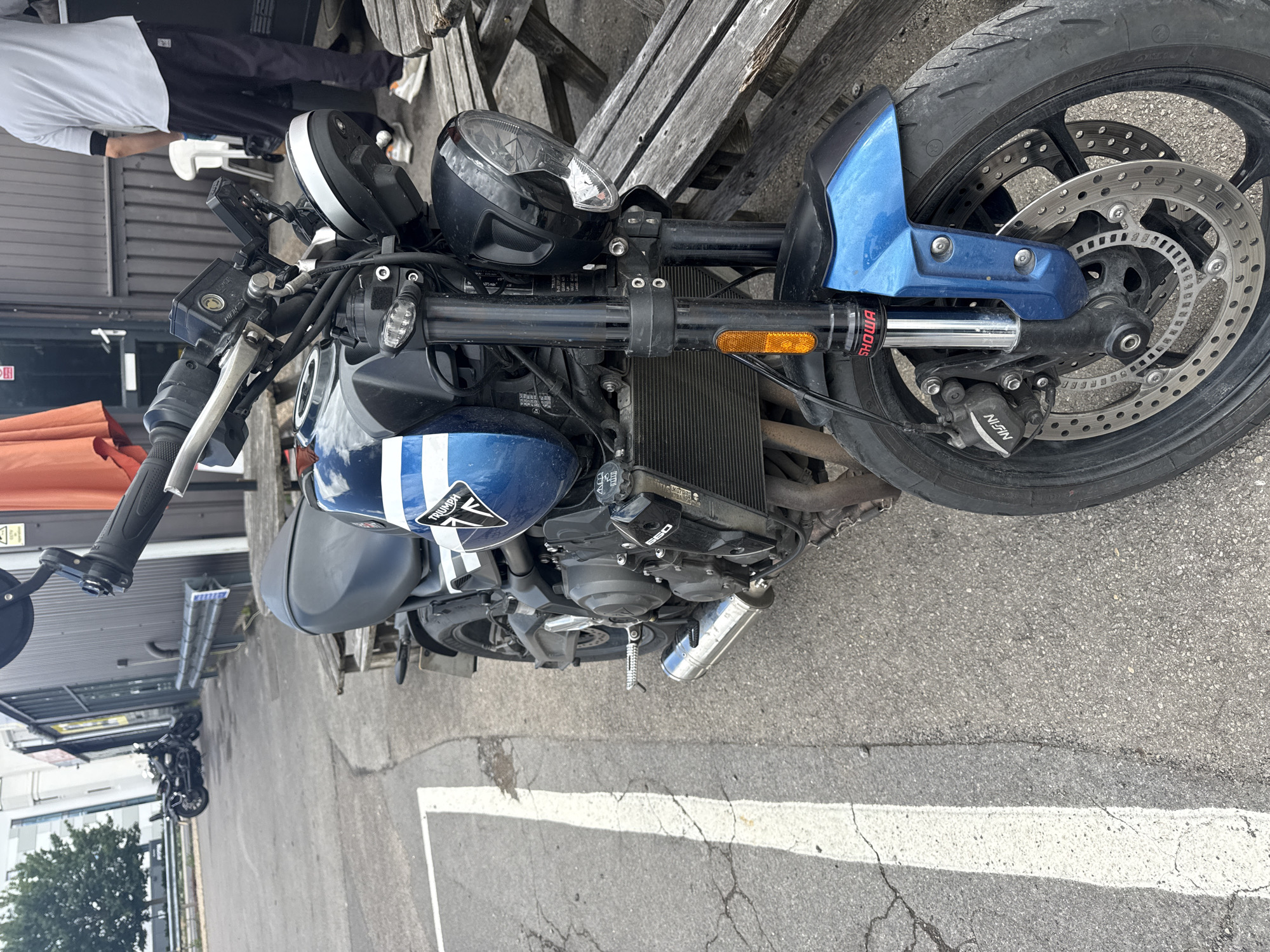 A blue and white Triumph motorcycle is parked on asphalt next to a wooden picnic table. The motorcycle is in the foreground, taking up most of the frame, with its front wheel, engine, and part of the fuel tank visible. The motorcycle has a black seat and handlebars, and chrome accents. In the background, a person wearing a grey t-shirt and black pants stands near a building with a grey corrugated metal facade. Two other motorcycles are parked in the distance, and a tree is visible to the left. The scene appears to be an outdoor area, possibly a cafe or rest stop, in Tewkesbury, United Kingdom.