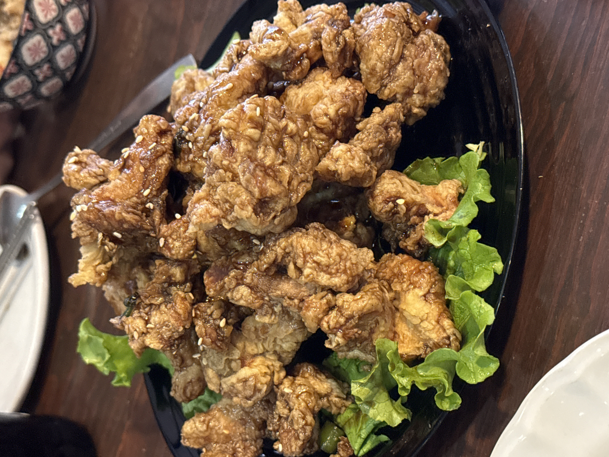 The image shows a platter of fried chicken pieces, garnished with green lettuce leaves. The chicken is golden-brown and appears to be coated in a glaze, with sesame seeds scattered on top. The platter is black and placed on a dark wooden table. In the periphery of the image, a bowl with a patterned design and silverware on a white plate are visible. The location is identified as Puebla, Mexico. There are no people visible in the image.