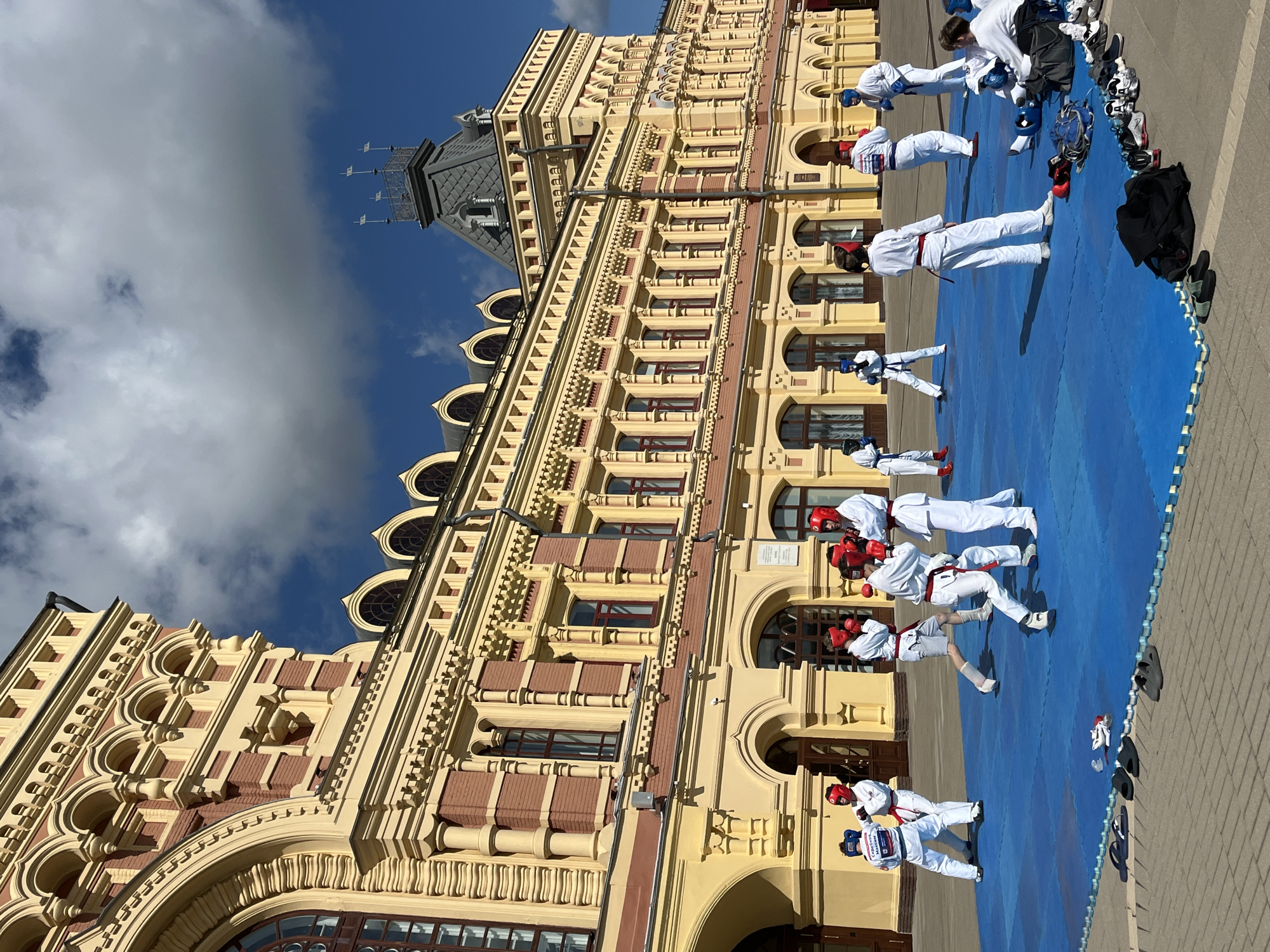 The image depicts an outdoor martial arts training session on a bright, sunny day in Nizhny Novgorod, Russia. A group of individuals, primarily young people, are practicing on a large blue mat in front of a grand, yellow and red building. They are dressed in white martial arts uniforms with red and blue protective gear, including helmets and gloves. Some are engaged in sparring, while others appear to be observing or waiting for their turn.
The architectural style of the building suggests it is a historical landmark, likely designed in a Neo-Russian or Renaissance Revival style, characterized by its intricate yellow ornamentation, arched windows, and red brick accents. The building dominates the background, its imposing facade creating a dramatic backdrop for the training activity. The sky is clear with scattered white clouds, indicating pleasant weather. On the ground around the mat, various athletic shoes and bags are placed, suggesting a break or preparation for training. The scene captures a moment of focused physical activity taking place in a public, urban space, likely a square or plaza adjacent to the historic building.