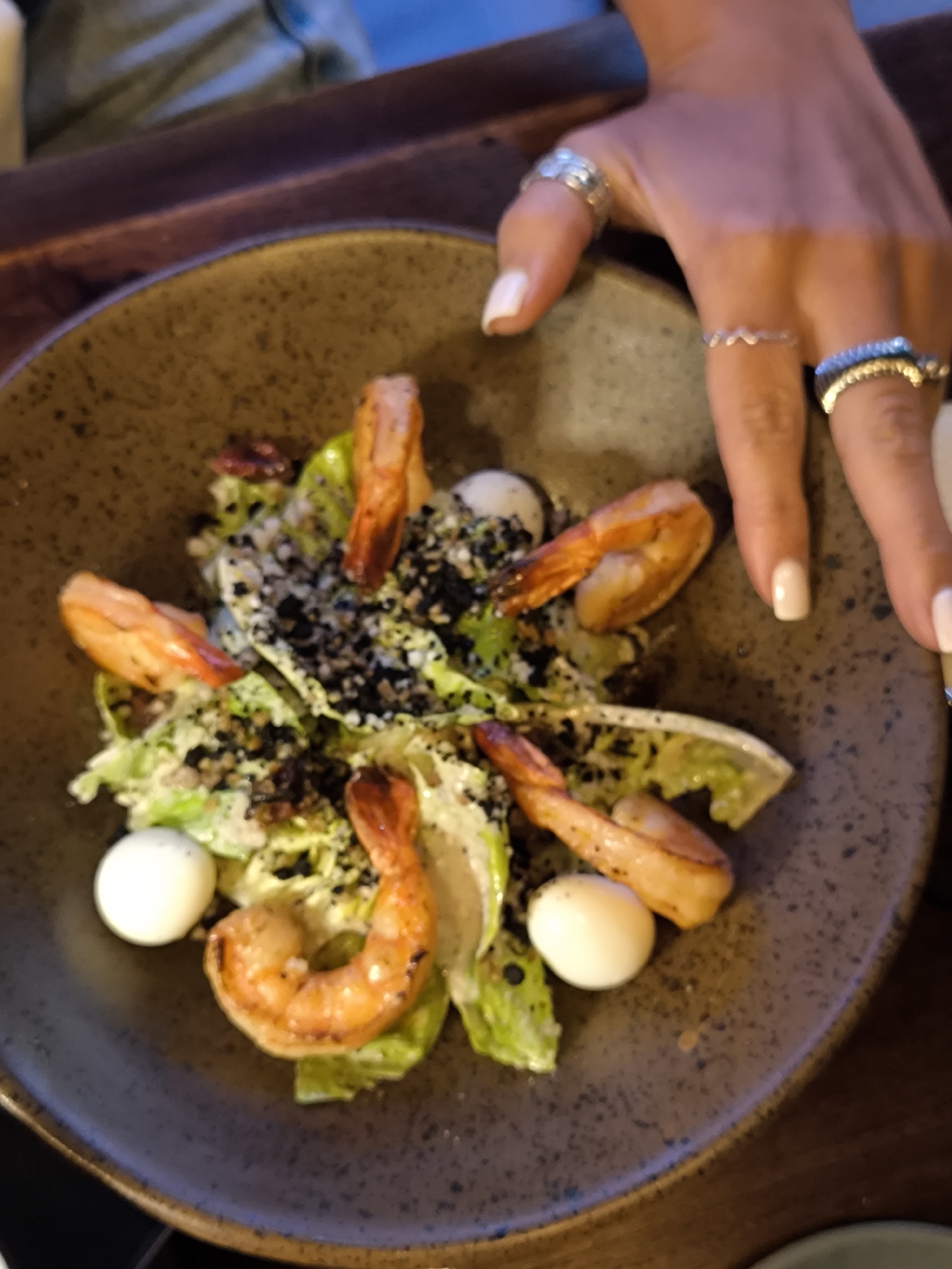 An overhead shot shows a dark ceramic bowl filled with a salad comprised of lettuce, grilled shrimp, hard-boiled quail eggs, and black, crumbly specks of an indeterminate substance. A person’s hand with multiple rings and light-colored polished nails is positioned next to the bowl. The bowl rests on a dark-toned wooden table. The scene suggests a dining establishment, possibly located in the Ubud District of Indonesia, based on the user prompt.