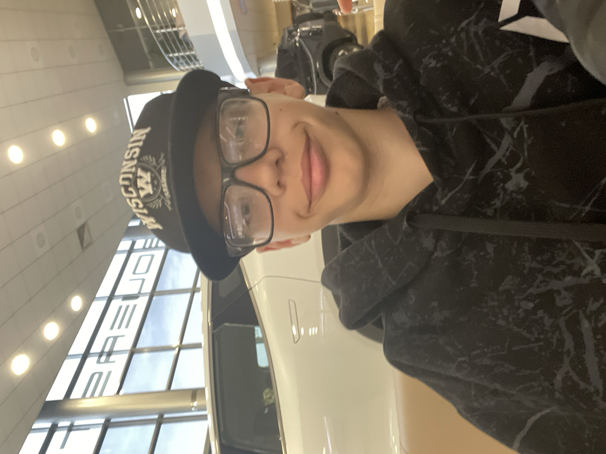 A young person wearing glasses and a black "WISCONSIN" baseball cap is taking a selfie in what appears to be a modern car dealership. The person is wearing a black hoodie with a marble-like pattern. Behind them, a white car is visible, and the dealership's interior features large windows and a tiered balcony with railings. The overall impression is a bright, indoor setting. There is no discernible information in the image to suggest a location in Yekaterinburg, Russia.