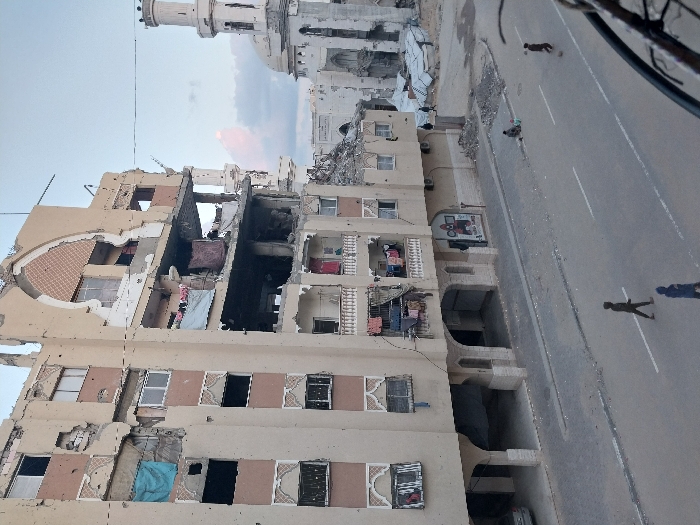 The image depicts a heavily damaged urban scene in Khan Yunis, Palestinian Territory, viewed from an elevated perspective.
In the foreground, a multi-story building with a light beige facade dominates the left and center. This building exhibits extensive structural damage, particularly on its upper and middle floors, where large sections of walls and ceilings are missing, revealing exposed interiors, rebar, and rubble. Decorative arched elements and window frames are visible, with many windows either shattered or boarded up with blue tarps. Laundry is hanging on a balcony on one of the lower damaged floors. Bullet or shrapnel marks are visible across the building's exterior.
To the right of this building, a heavily damaged mosque with a minaret stands. Its dome and main structure are significantly compromised, appearing as a skeletal shell. Piles of rubble are visible on the ground between the main building and the mosque.
At street level, a paved road with white dividing lines stretches across the lower portion of the image. Four individuals are present on the road: one person in dark clothing walks left, followed by another in a blue hooded garment. Further down, a person in dark clothing is interacting with a ball, and another figure stands near them. On the sidewalk adjacent to the main building, two individuals are seated near a small pile of rubble. A light-colored car is partially visible, parked under an archway on the ground floor of the main building.
The sky above is light blue with scattered clouds, one showing a hint of pink. A dark railing or balcony edge is visible in the bottom right corner, suggesting the vantage point of the photograph.