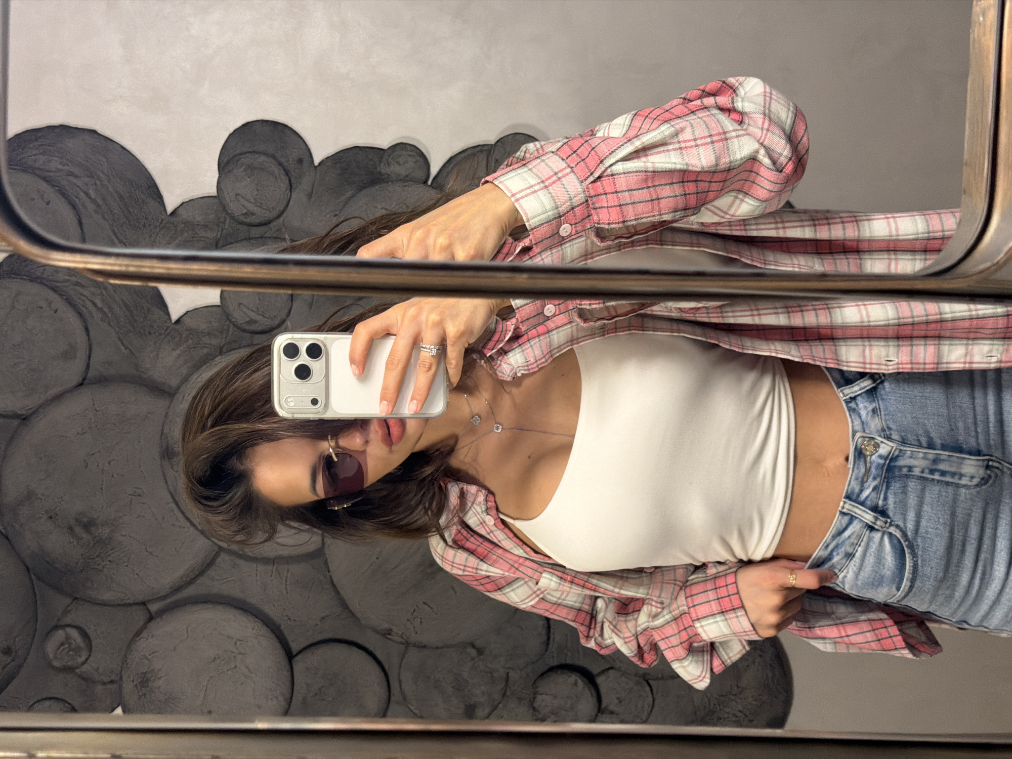 A young woman takes a selfie in a mirror, likely in a restroom or changing room. She is fair-skinned with brown hair, wearing sunglasses, a white tank top, a pink and white plaid shirt (unbuttoned), and light-wash blue jeans. She holds a white smartphone in her right hand. The background shows a textured wall with large, rounded shapes that resemble stones or bubbles. The mirror has a dark metal frame. The image appears to be taken indoors, but the exact location in Krasnoyarsk, Russia, cannot be determined from the visual information alone. There is no indication of the location's specific coordinates or address visible in the image.