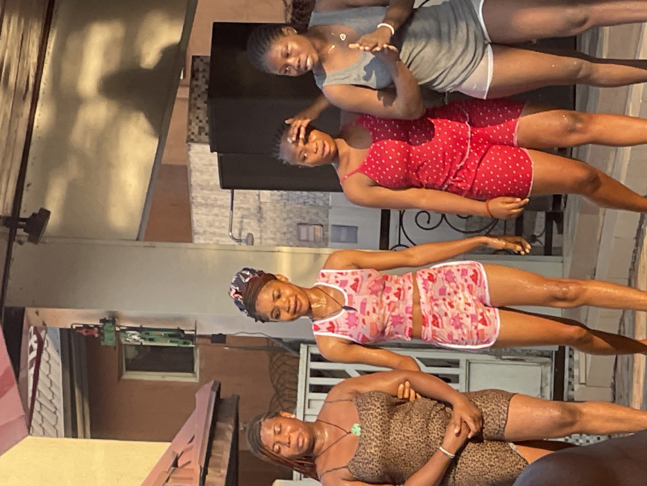 Four young women pose on what appears to be a balcony. One wears a gray tank top and shorts, while another wears a red polka-dot outfit. A third is in pink and white patterned pajamas, and the fourth dons a leopard-print outfit. All seem to be sweaty and are smiling. The background includes building facades and fencing.