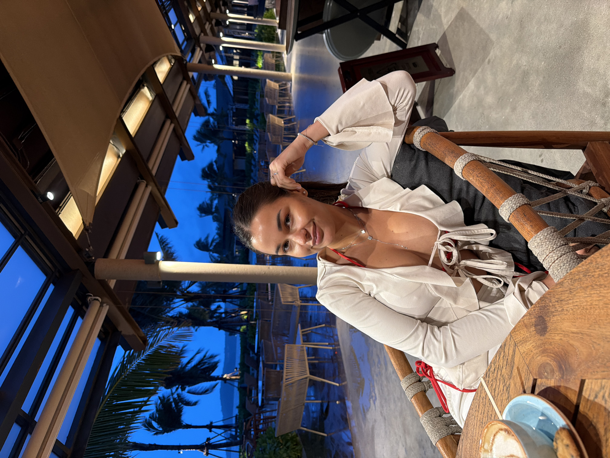 A seated woman is captured in an indoor setting at Choeng Thale, Thailand. The woman is smiling. She wears a white top over a red garment. A beverage and blue plate are visible on the wooden table, adjacent to the woman. The surrounding environment includes outdoor seating, palm trees, and a view of the ocean at dusk. The interior has an open design with structural beams and soft lighting.