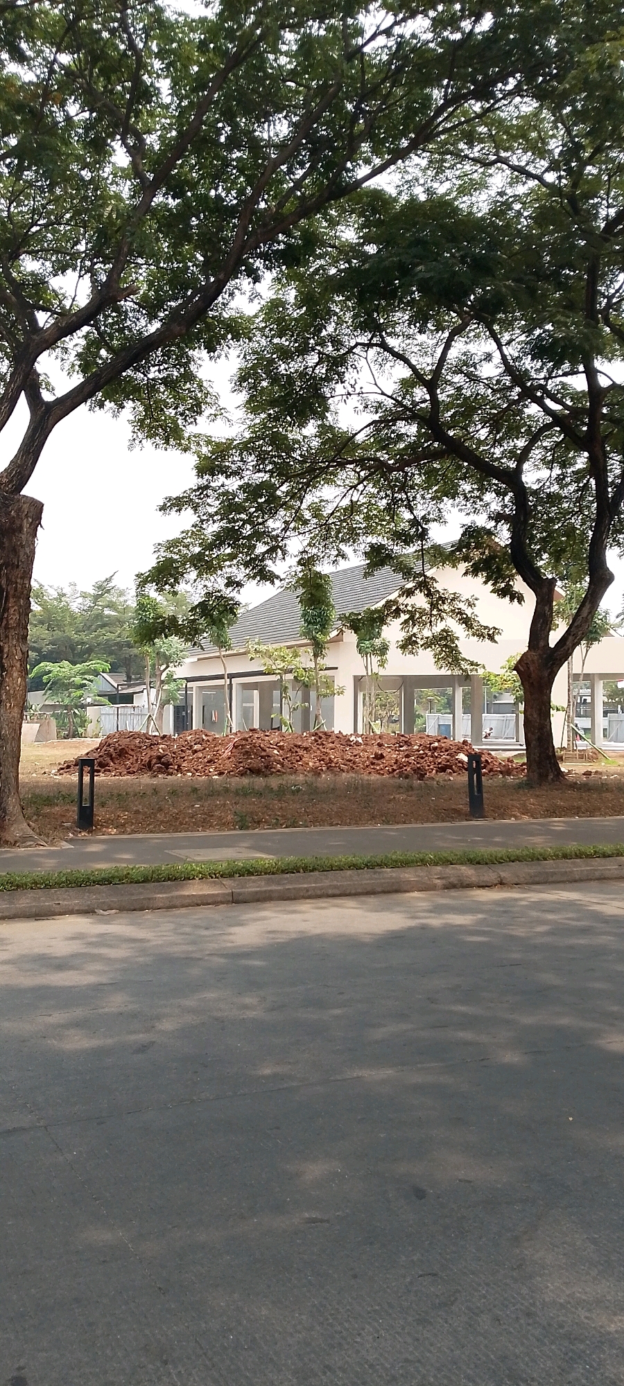 The image depicts a construction site with a partially built modern building in the background. The building has a gray tiled roof and large glass-paneled openings, suggesting it may be a commercial or residential complex. In front of the building, there is a large mound of reddish-brown soil, indicating ongoing landscaping or construction activity. Two dark, rectangular outdoor lights are visible in the foreground, placed on either side of the soil mound. The scene is framed by the branches and leaves of large trees, casting dappled shadows on the asphalt road in the lower part of the image. A narrow strip of green grass separates the road from the construction area. The sky is overcast and bright, suggesting a daytime scene. The location is identified as Bekasi, Indonesia.